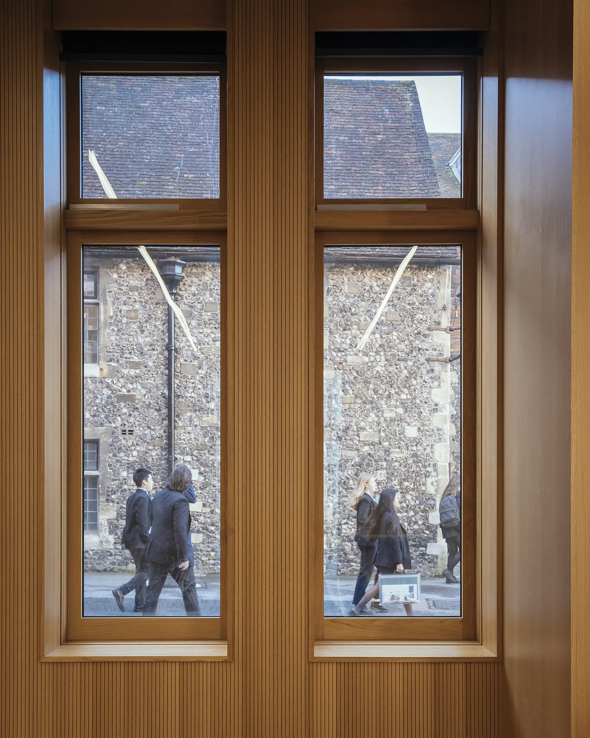 Interior view through timber-framed windows showing pedestrians passing weathered stone walls outside