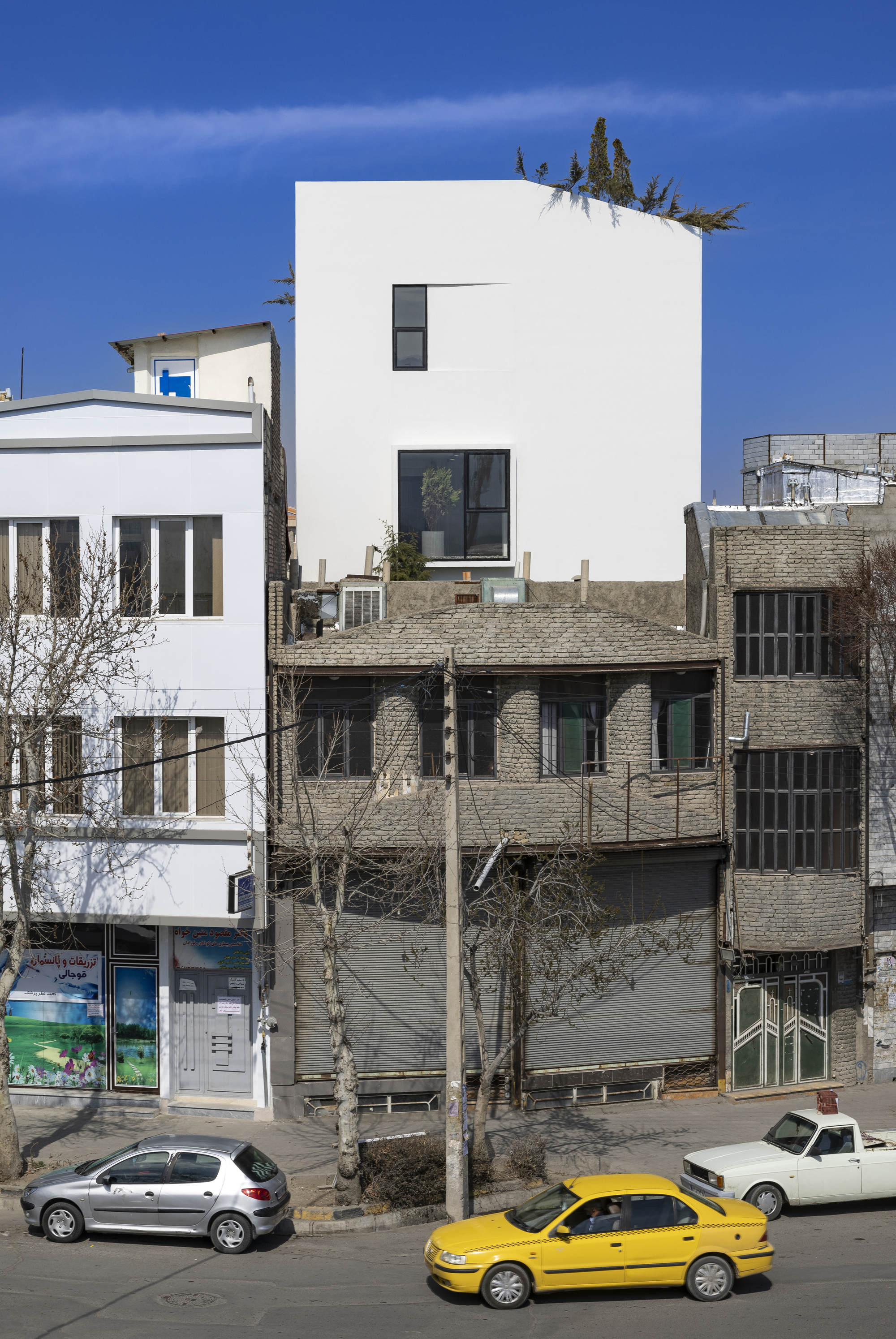 Street view of a white cubic volume rising above neighboring brick structures with a yellow taxi passing