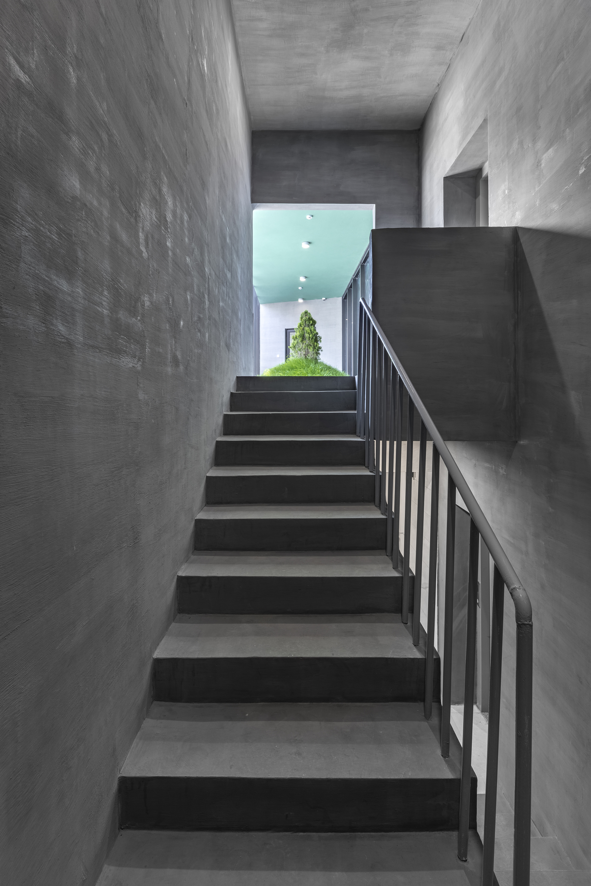 Concrete staircase with metal railing ascending toward a turquoise ceiling and glimpse of greenery