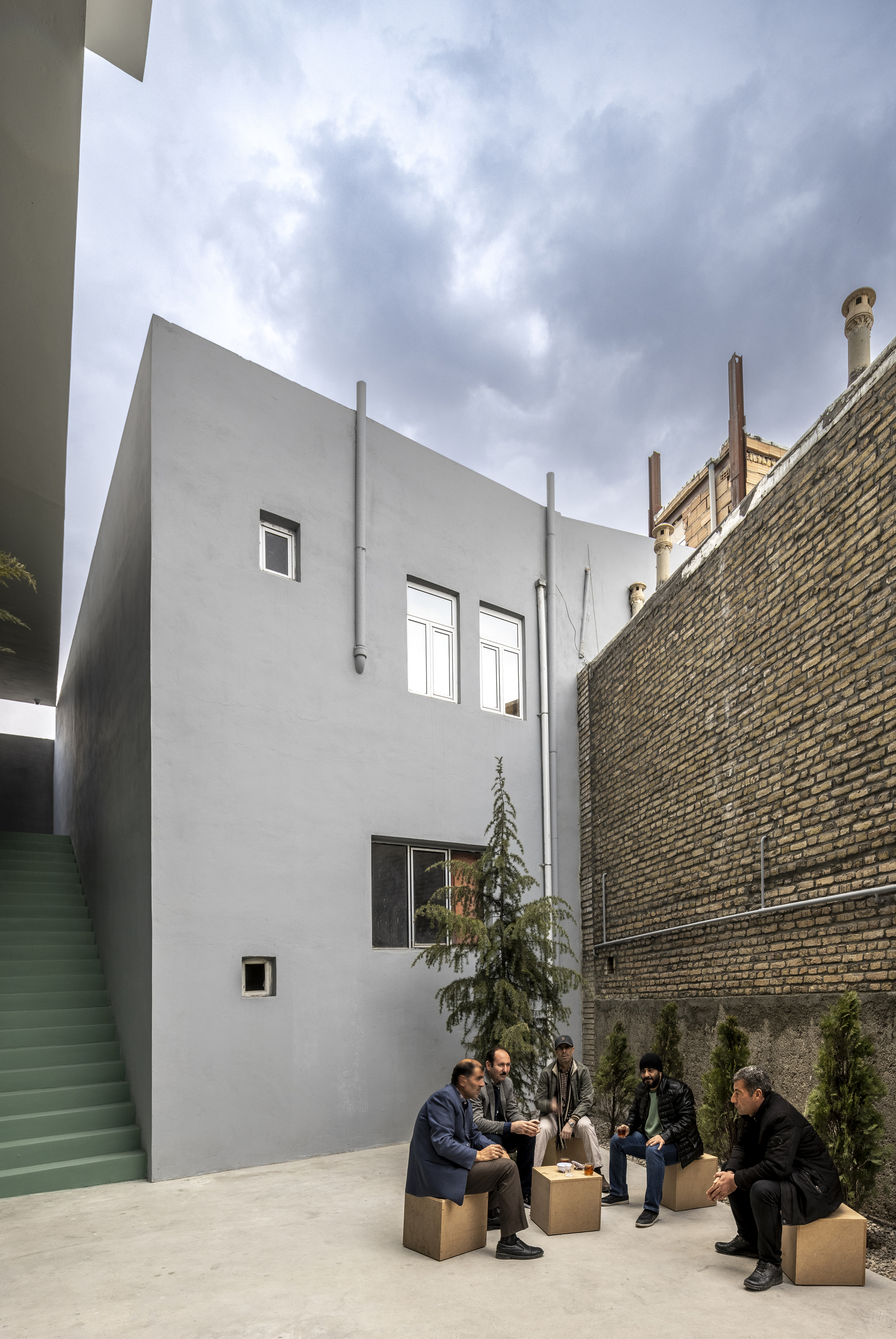 Courtyard view with white rendered facade, brick party wall and people gathered on cardboard seating
