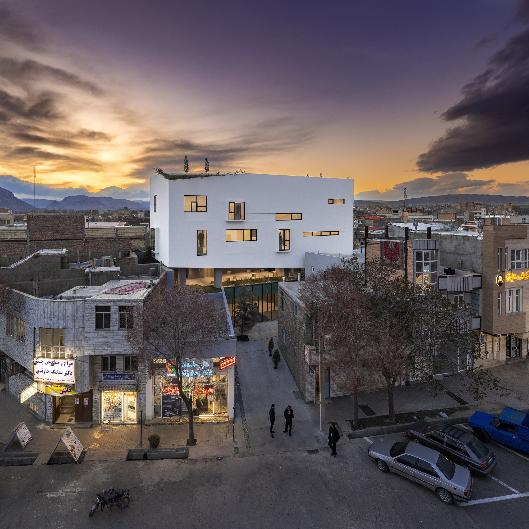 White volume with illuminated windows at dusk above pedestrians and storefronts in the street below