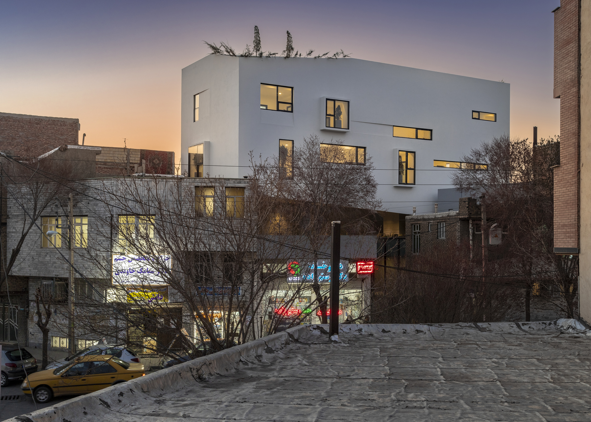 White facade with scattered window openings lit from within at twilight over bare winter trees