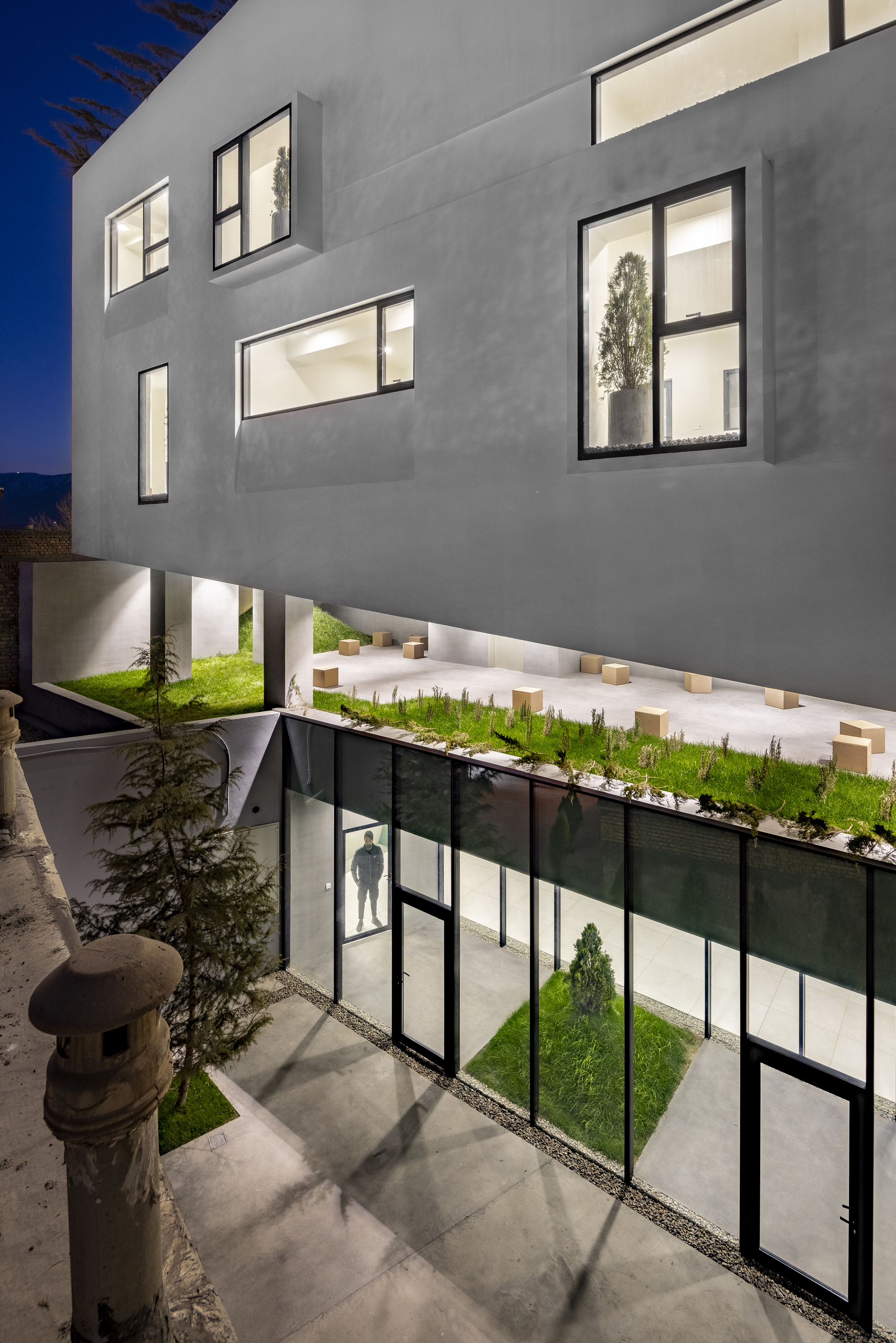 Planted courtyard with glazed walls below cantilevered white volumes at blue hour