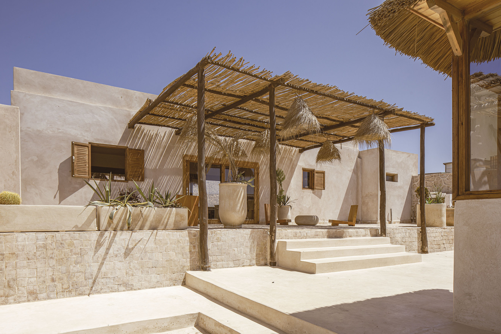 Exterior terrace with timber pergola and thatched roof over stone planters and steps in bright sunlight