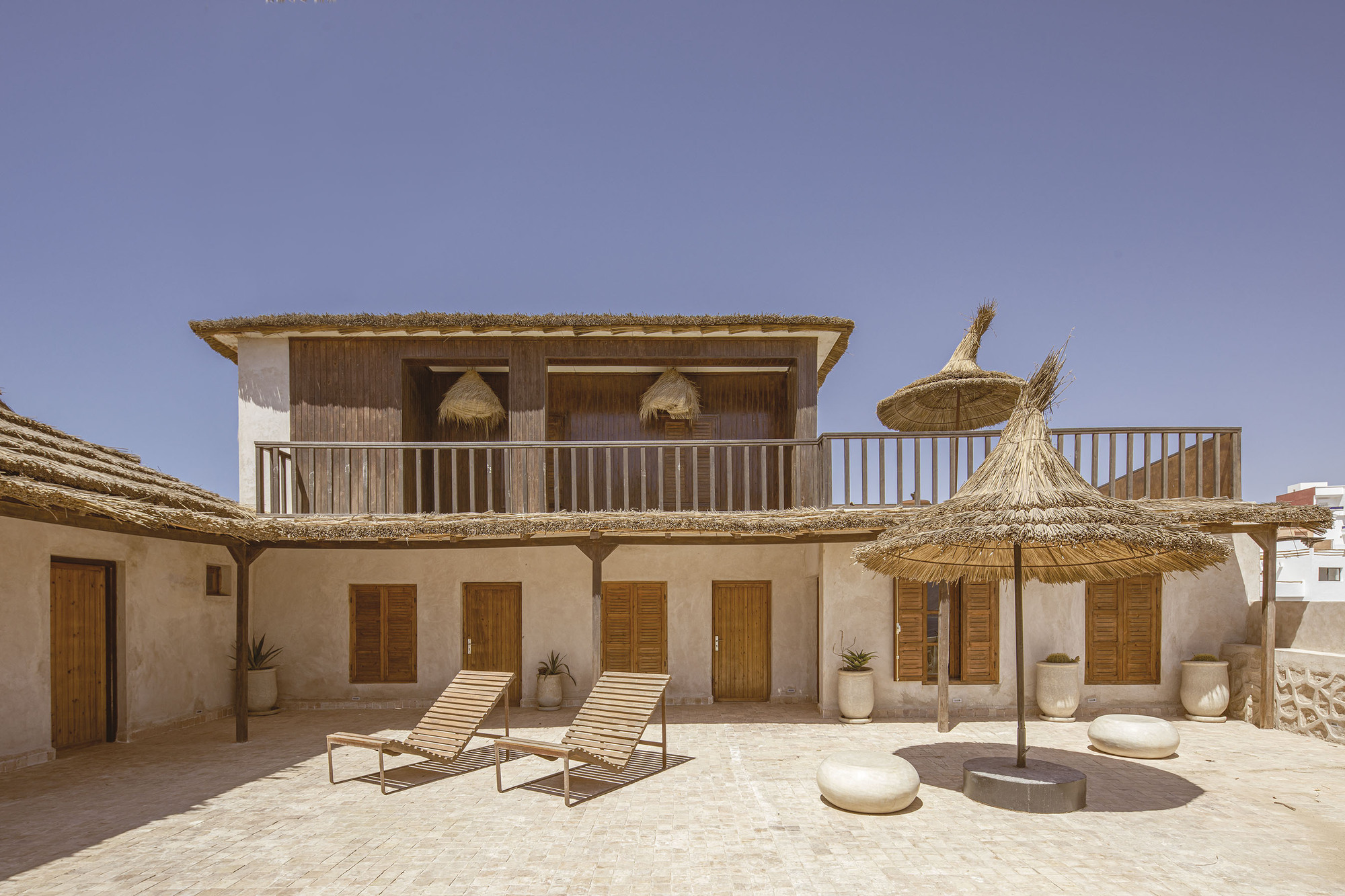 Two-story facade with timber shutters and upper balcony overlooking terrace with lounge chairs and thatch umbrellas