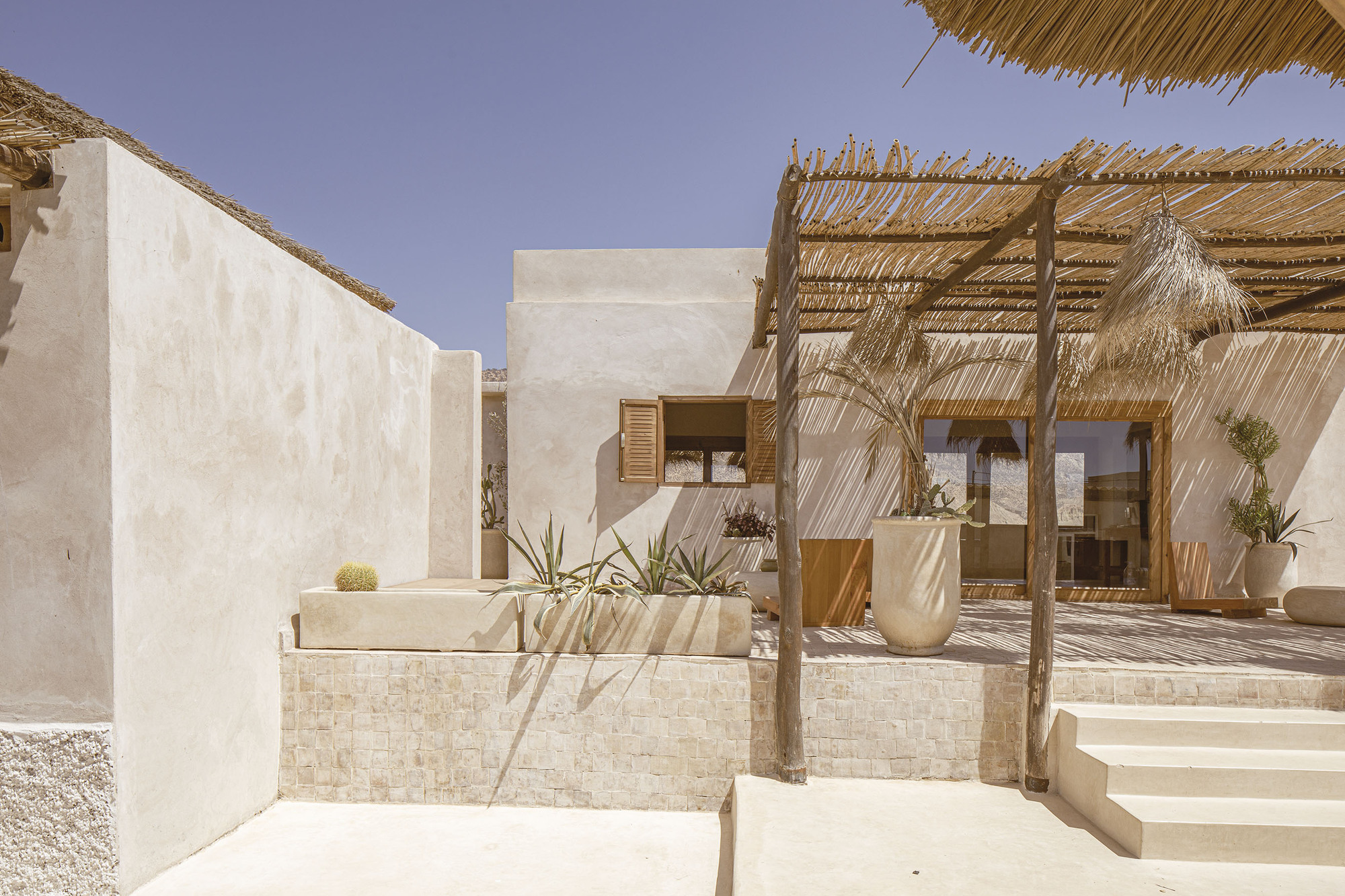 Exterior courtyard with rendered walls, stone planters, timber pergola with palm frond shade, and desert plants in bright sunlight