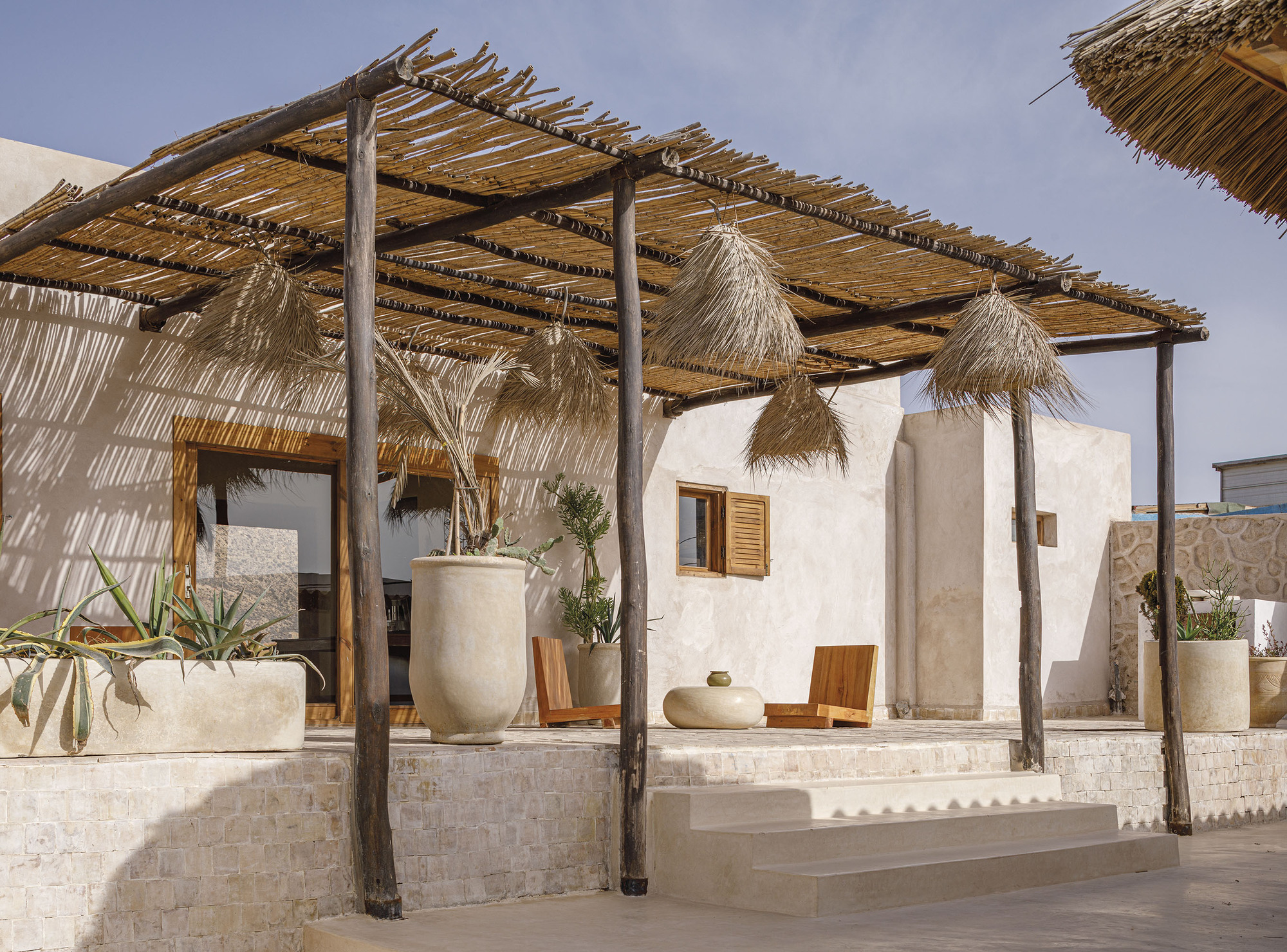 Outdoor pergola structure with bundled reed canopy, timber columns, hanging thatch shades, and planted ceramic vessels on stone platform