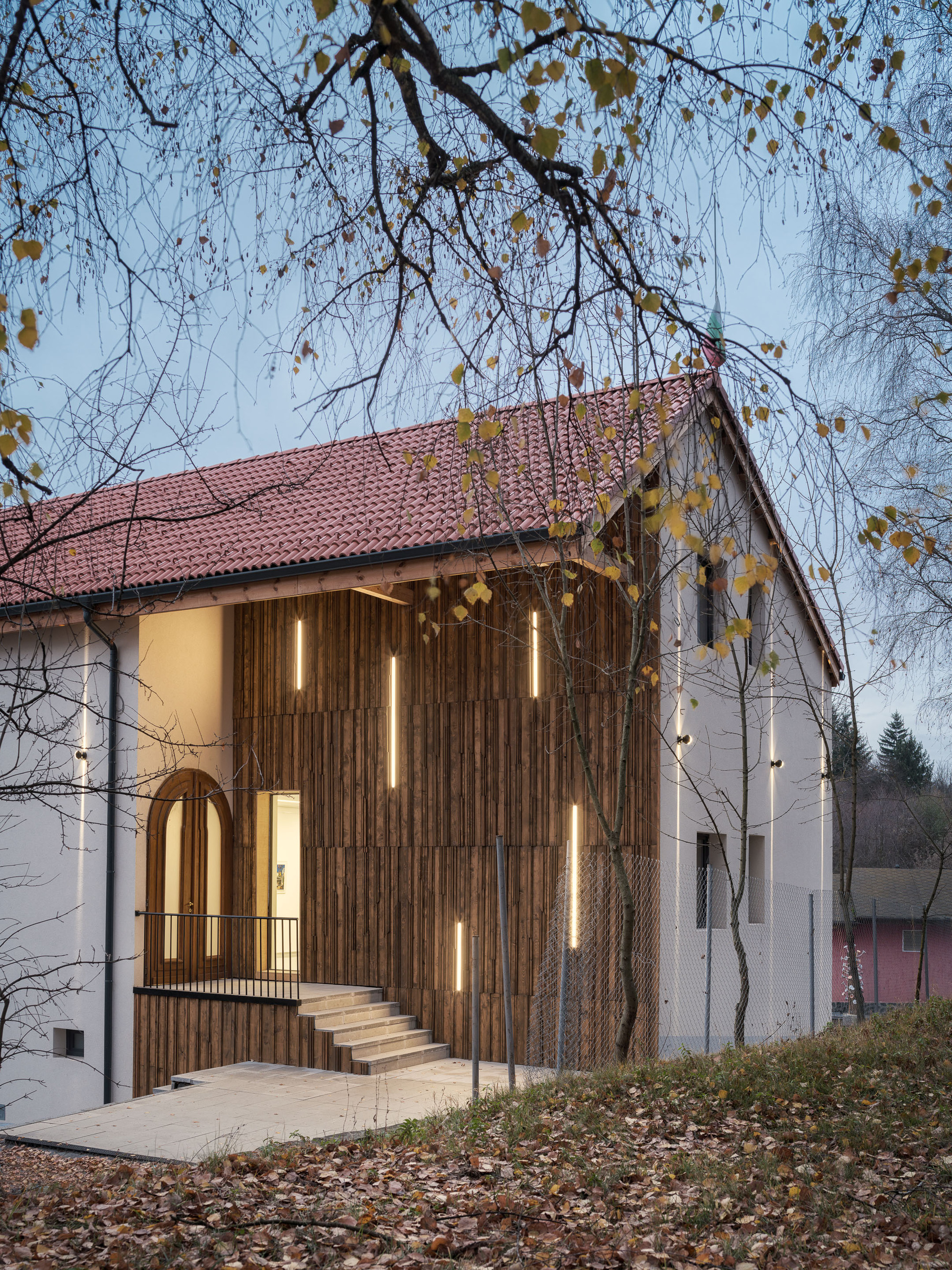 Timber-clad entrance facade with illuminated slat openings beneath bare tree branches in autumn
