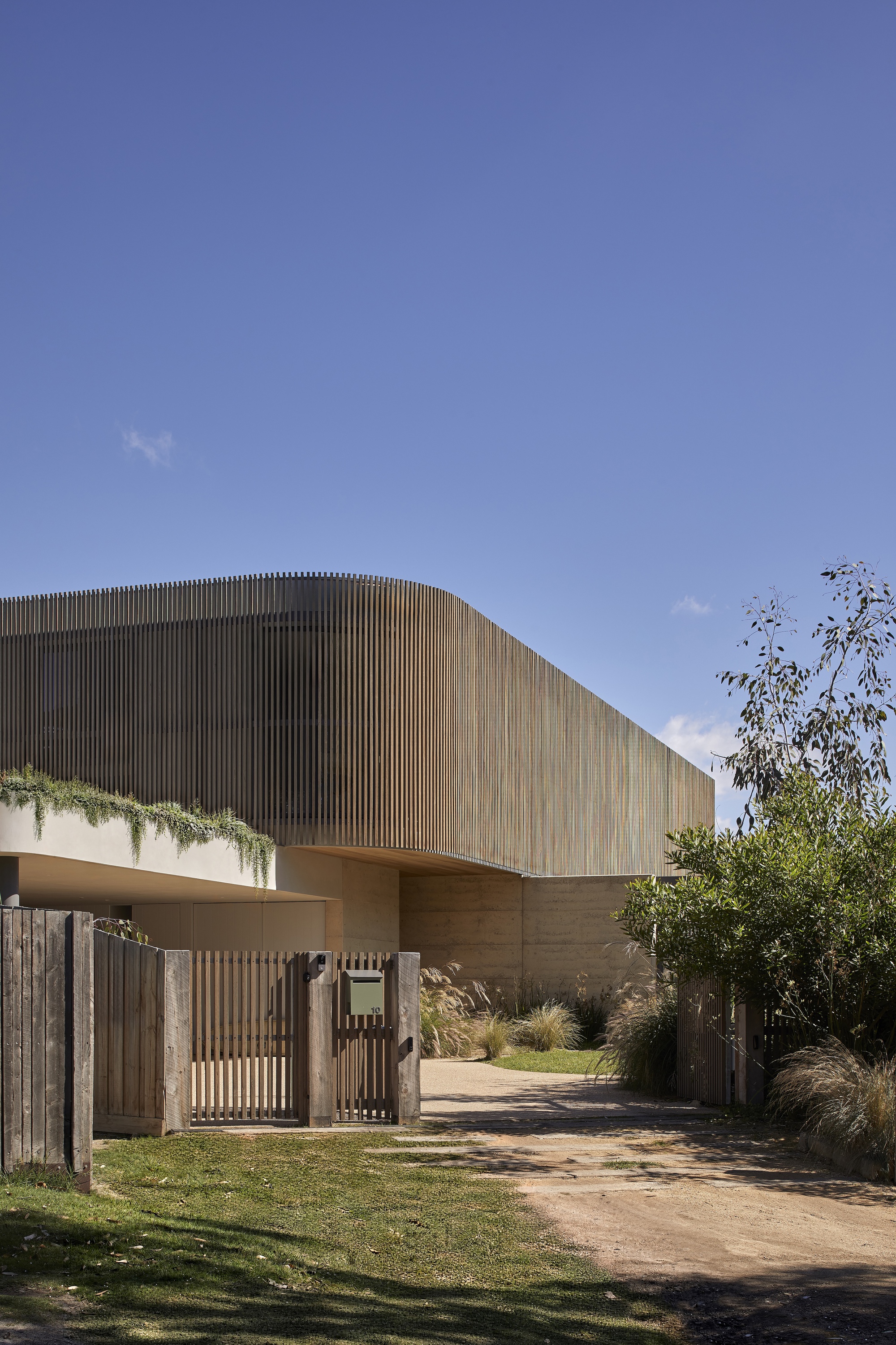 Curved timber-clad upper volume above rammed earth base with slatted gate and lawn