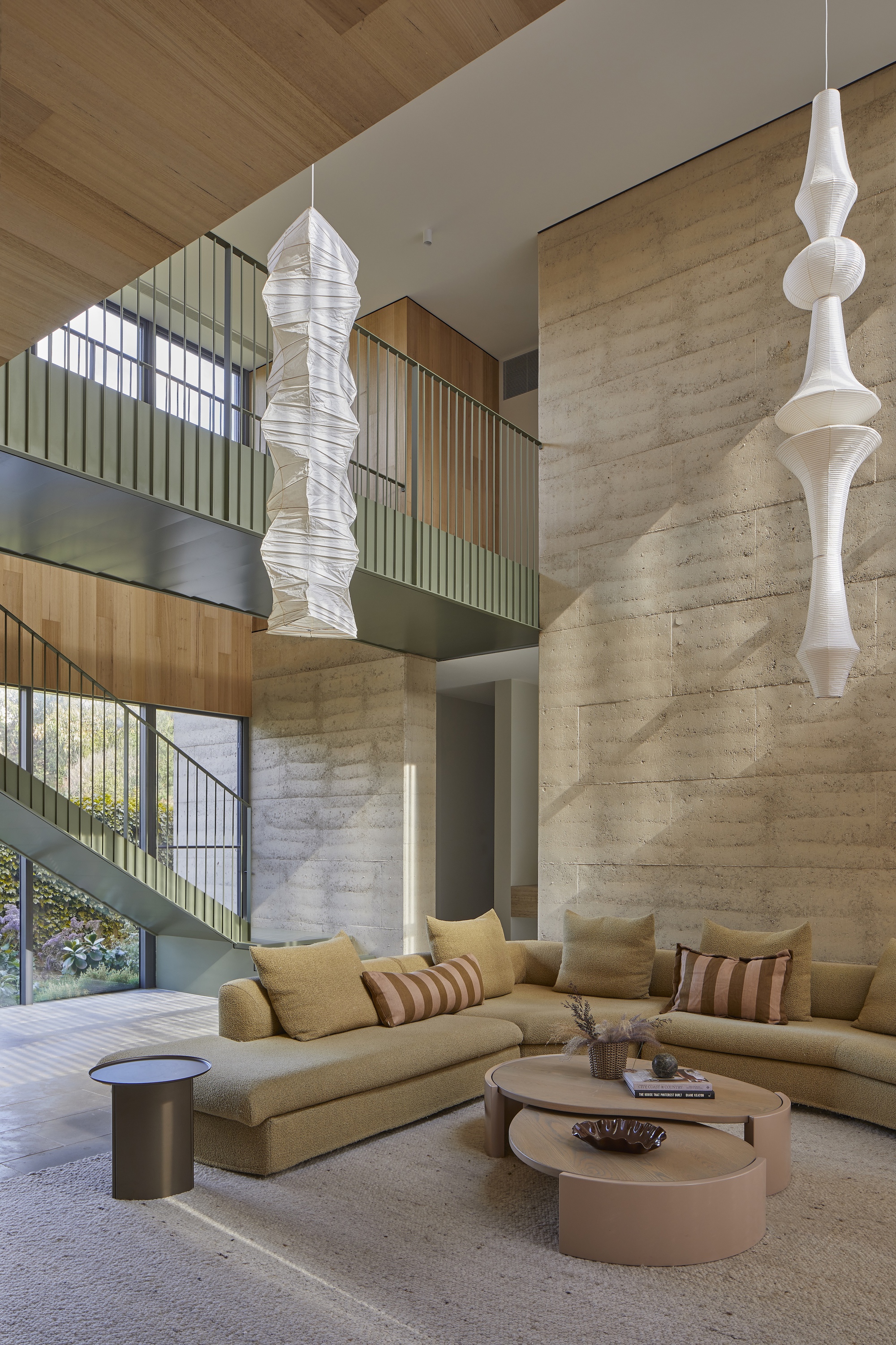 Double-height living space with rammed earth walls, suspended pendants and timber-clad balcony