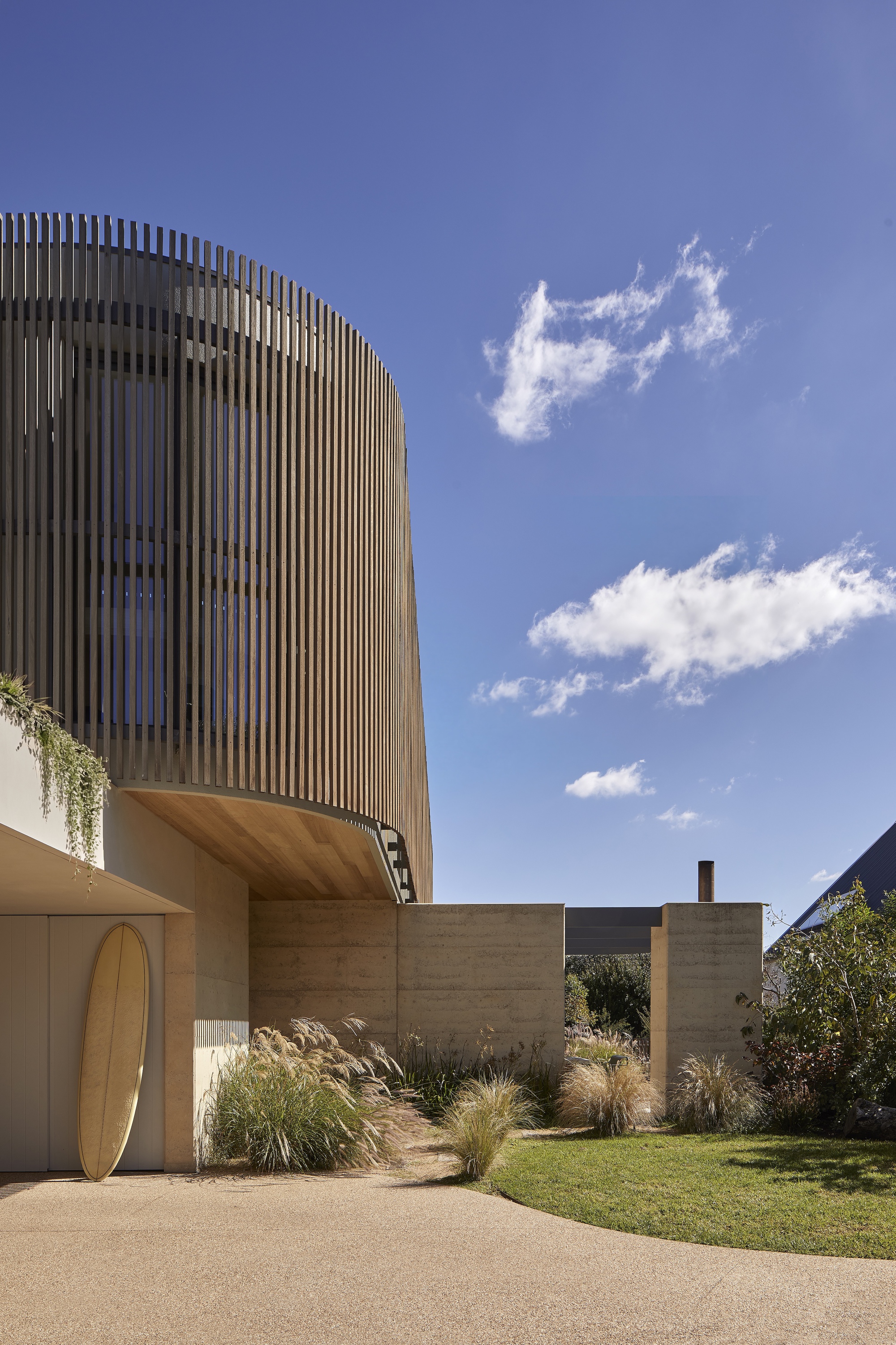 Two-storey facade with vertical timber screening above rammed earth walls and planted garden beds