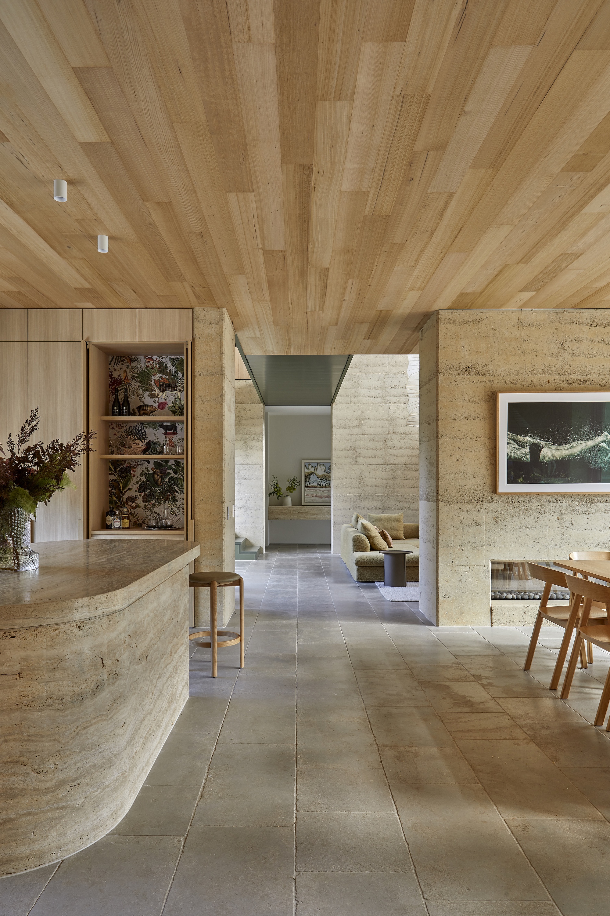 Open plan living space with timber plank ceiling and curved rammed earth kitchen island