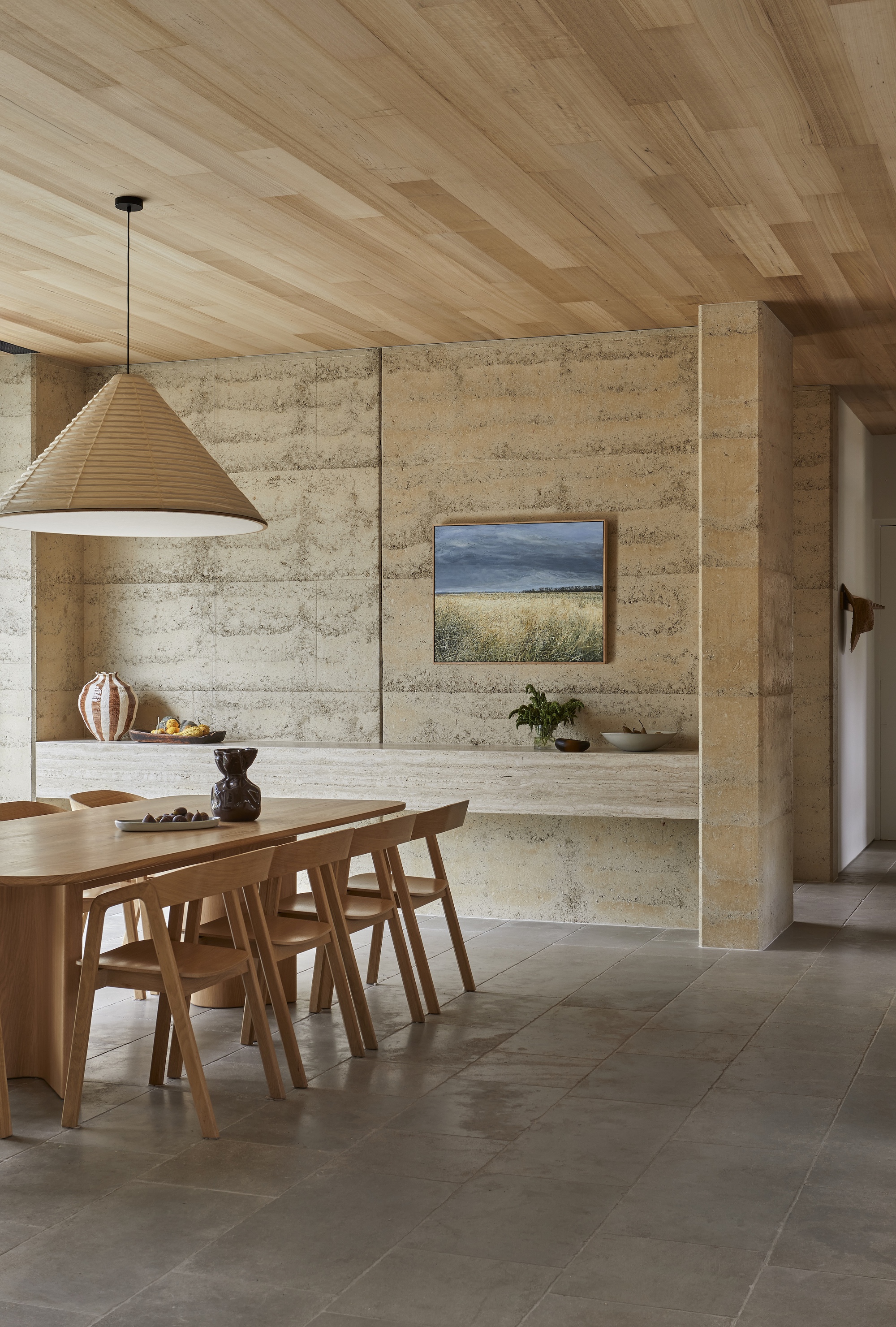 Dining area with rammed earth walls, timber plank ceiling and large pendant light above an oval table