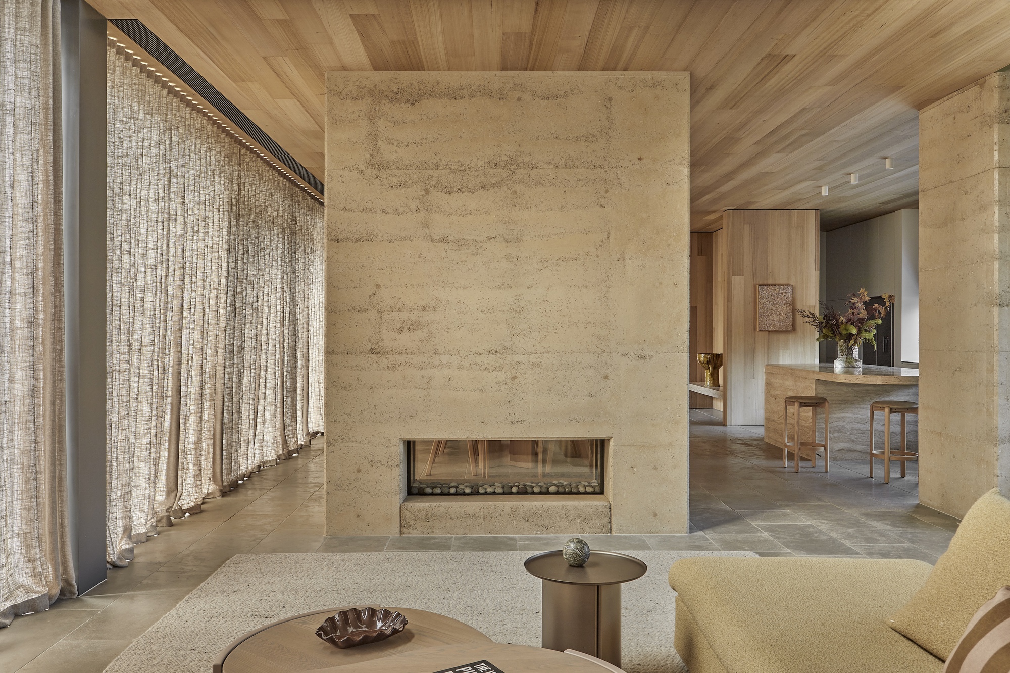 Rammed earth fireplace wall with linen curtains and view through to dining area beyond