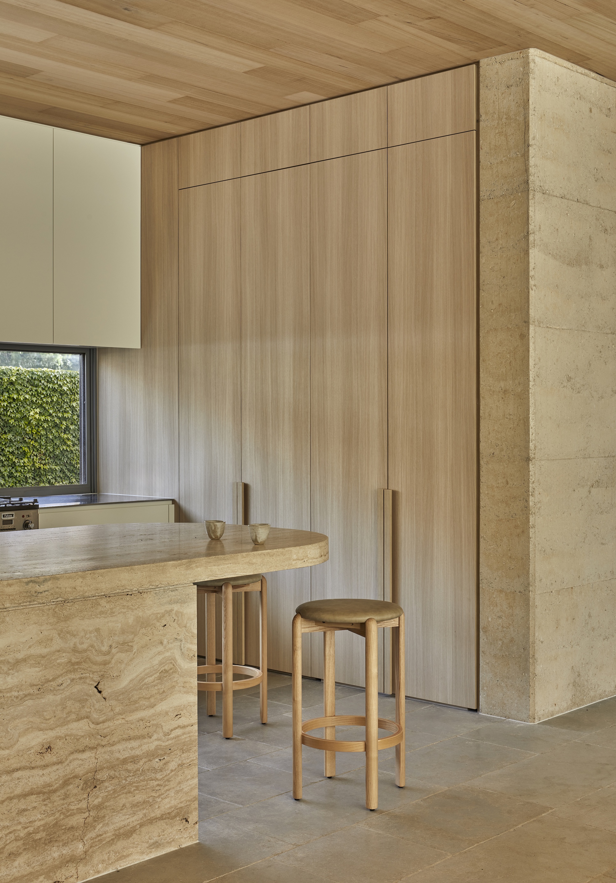 Kitchen with vertical grain timber cabinetry beside a rammed earth breakfast bar and two stools