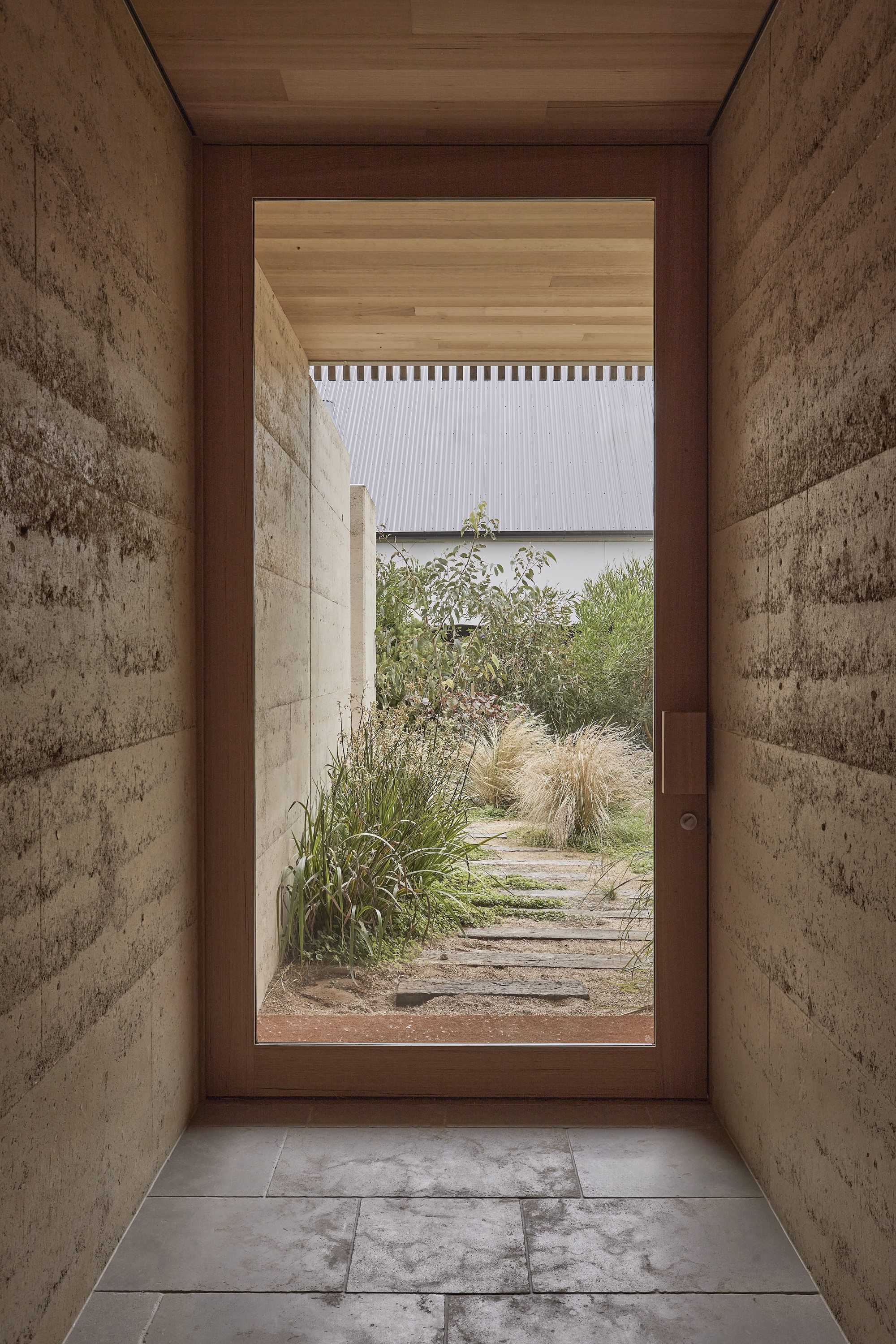 Glazed timber door framed by rammed earth walls opening to a gravel path and grasses