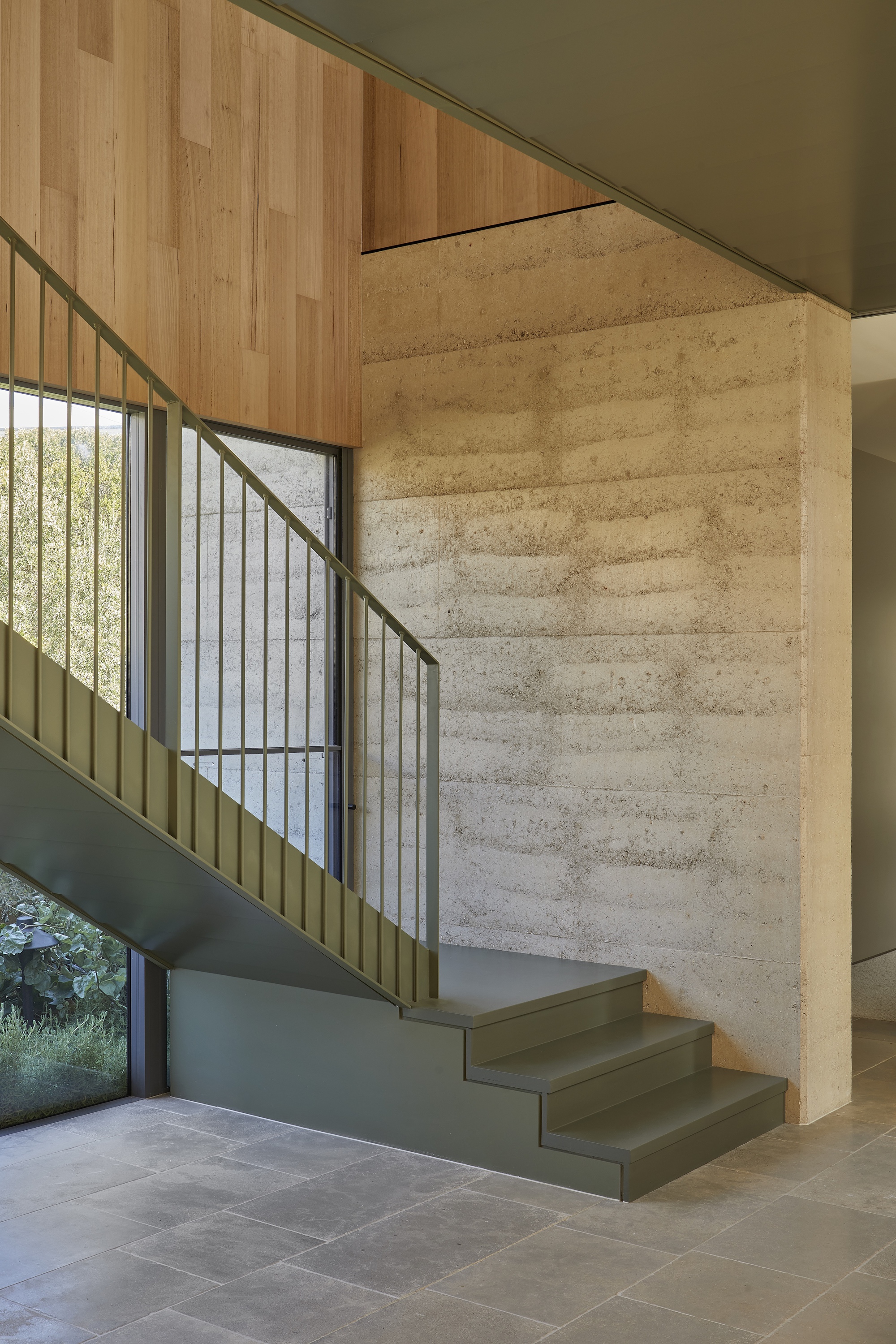 Staircase with metal balustrade rising beside a rammed earth wall and timber ceiling above