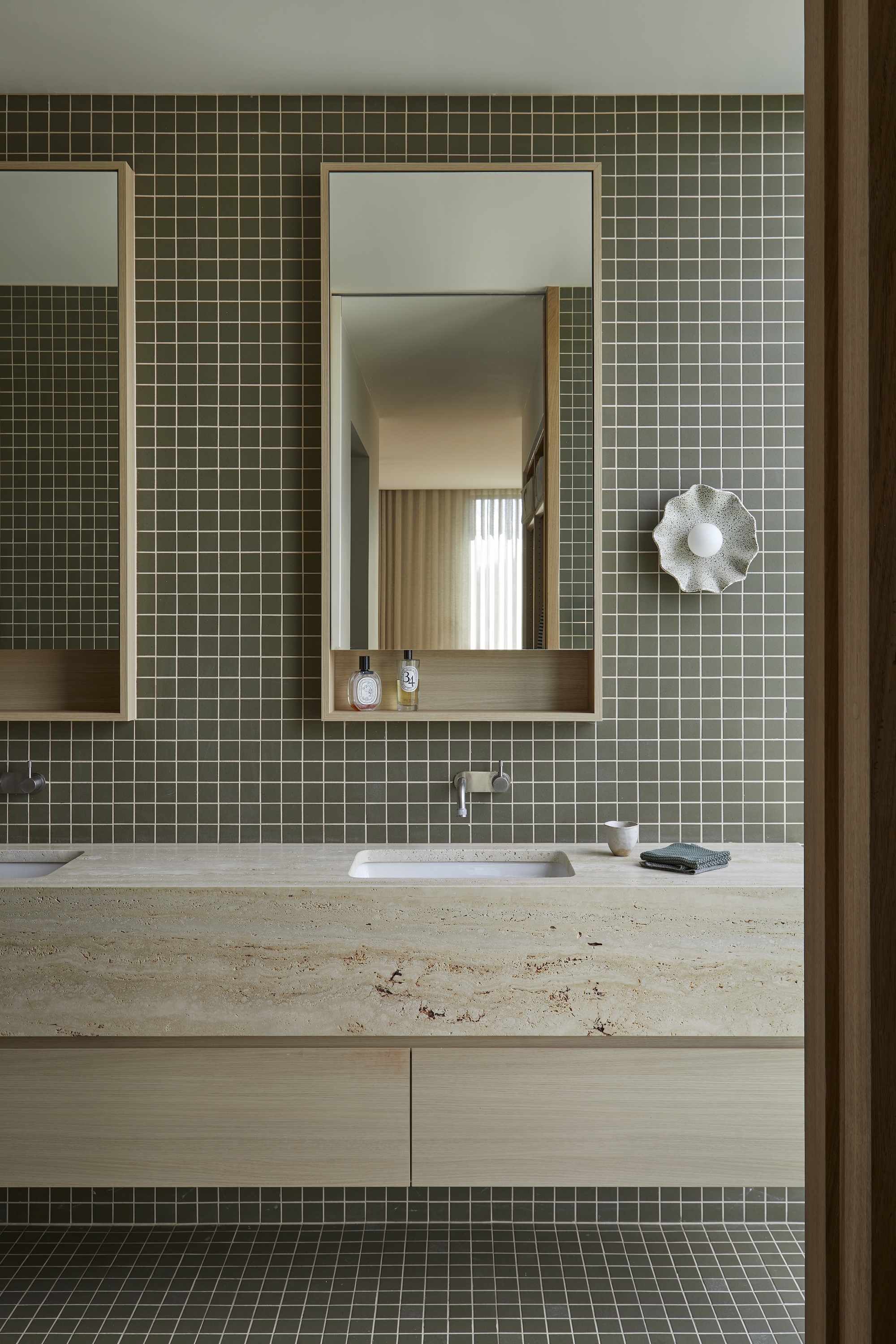 Bathroom vanity with olive green mosaic tile and travertine countertop below framed mirrors
