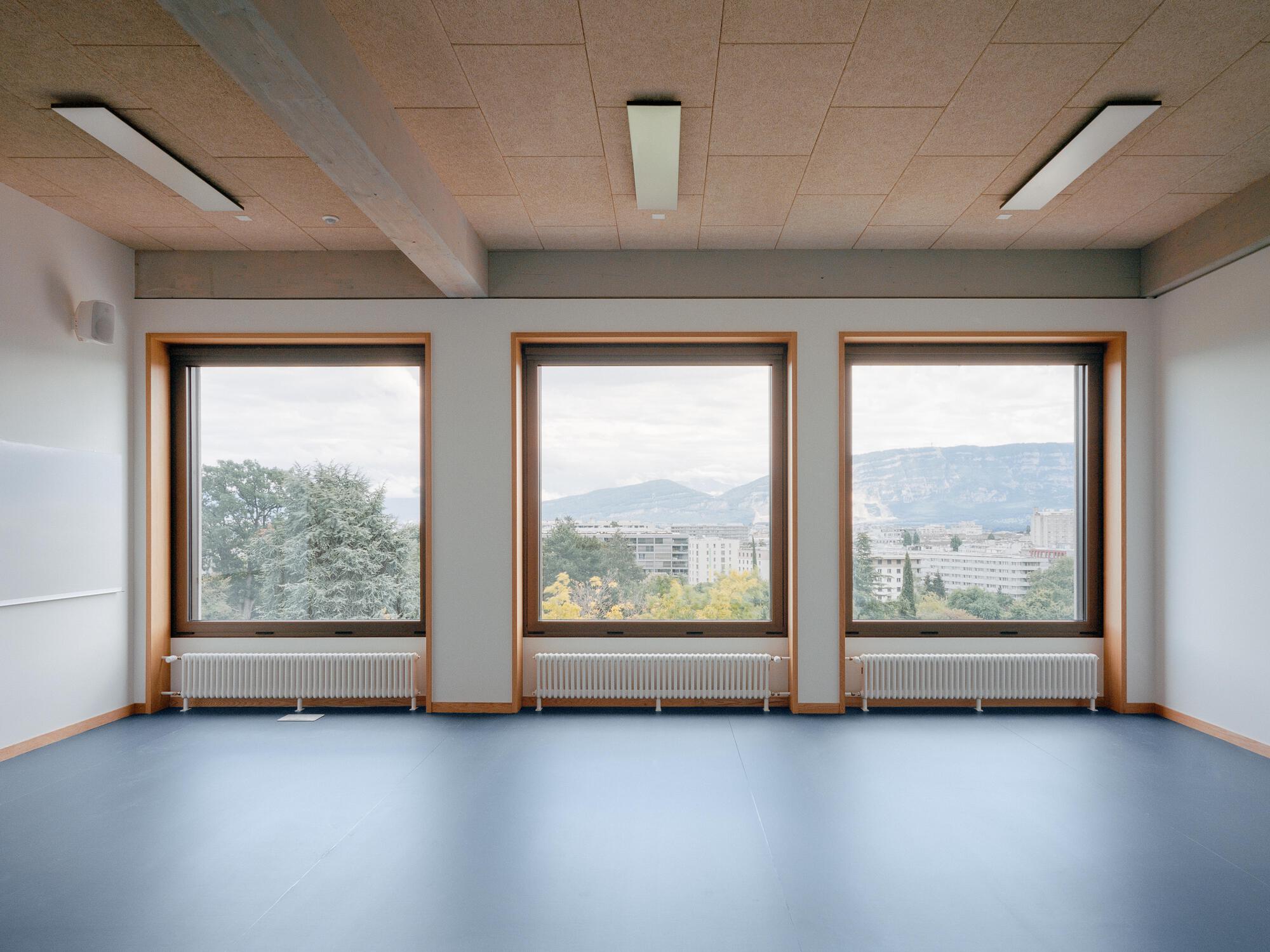 Empty room with three timber-framed windows overlooking distant mountains and residential blocks in overcast weather