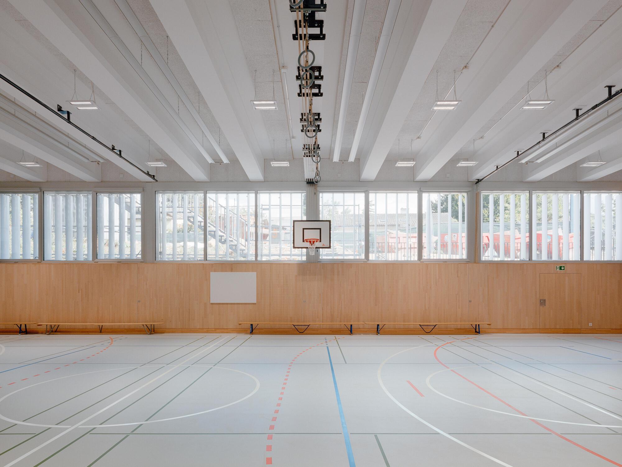 Gymnasium with painted floor lines, basketball hoop, exposed white ceiling beams and translucent perimeter screens