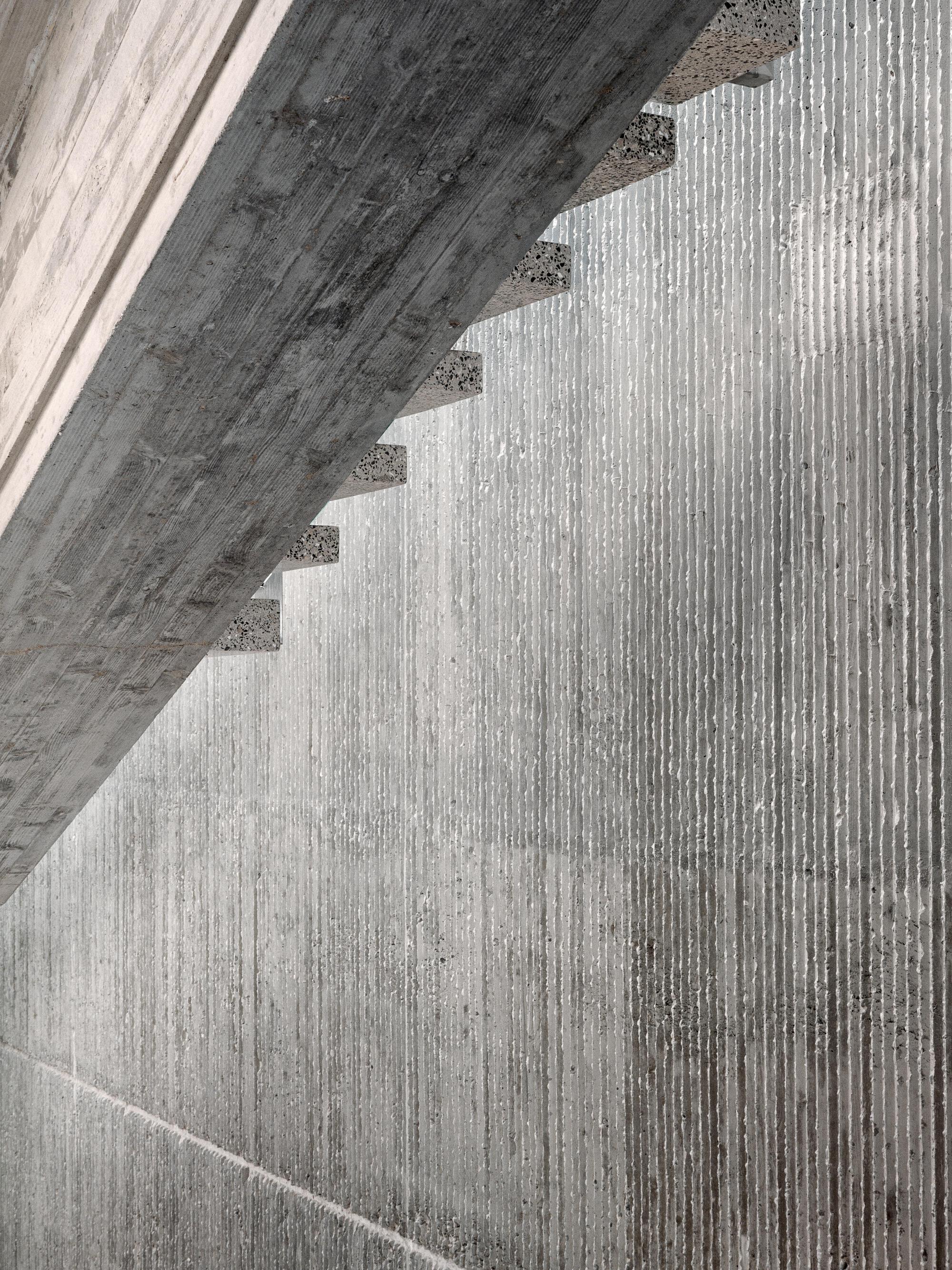 Close-up of board-formed concrete stair soffit casting diagonal shadows across ribbed wall