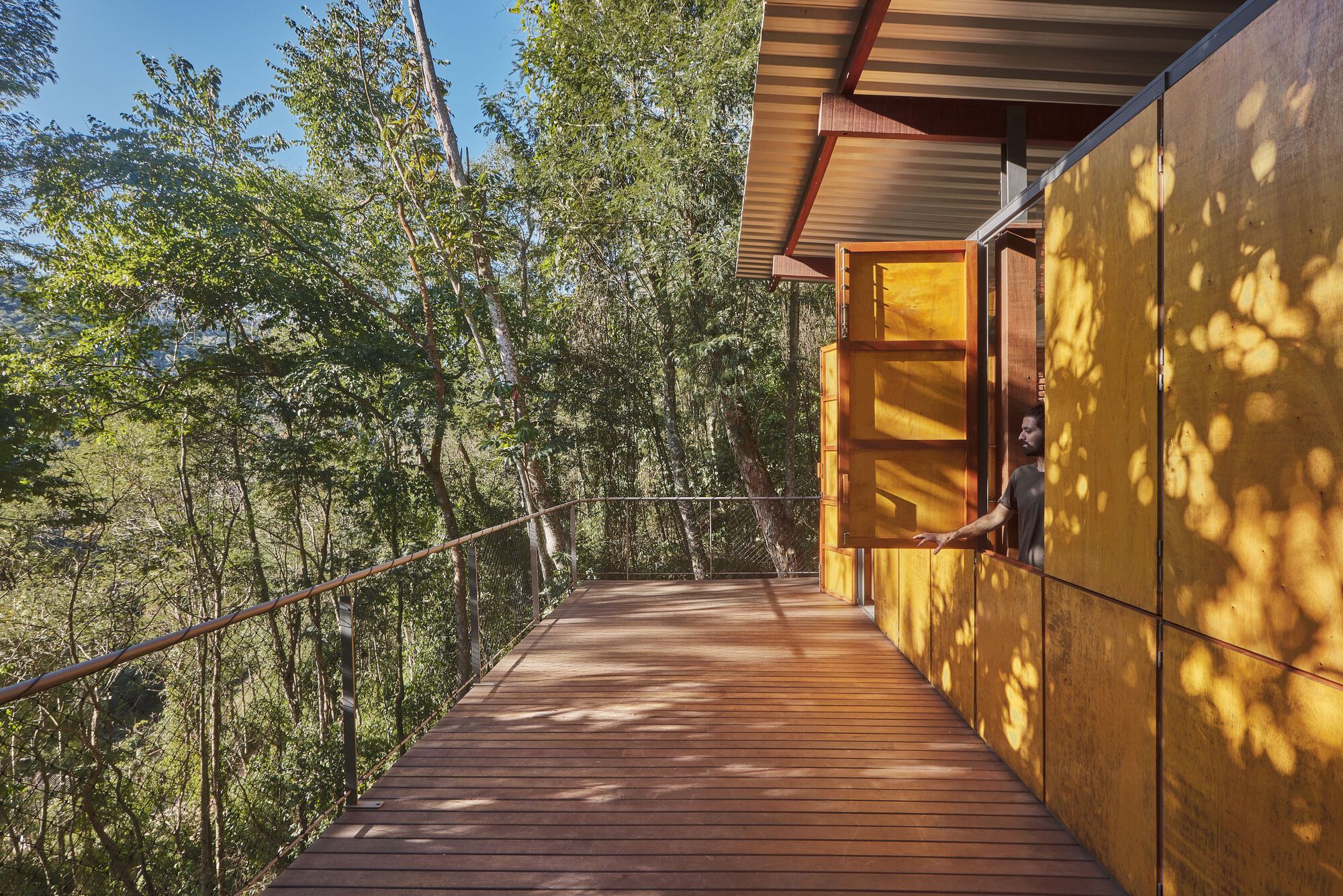 Timber deck with steel cable railing extending from yellow paneled facade with dappled sunlight