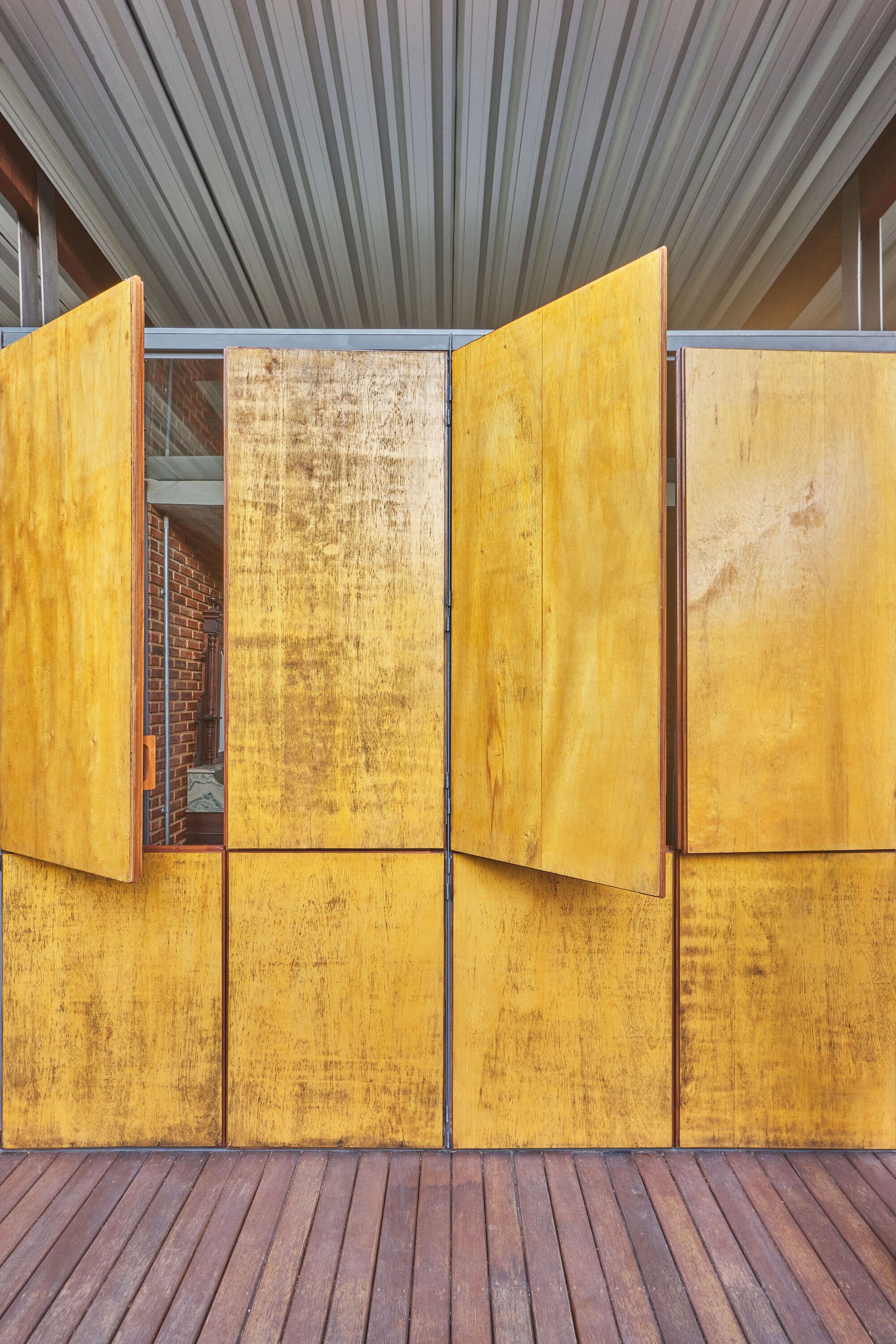 Weathered yellow plywood facade with operable shutters beneath a ribbed metal overhang and timber decking