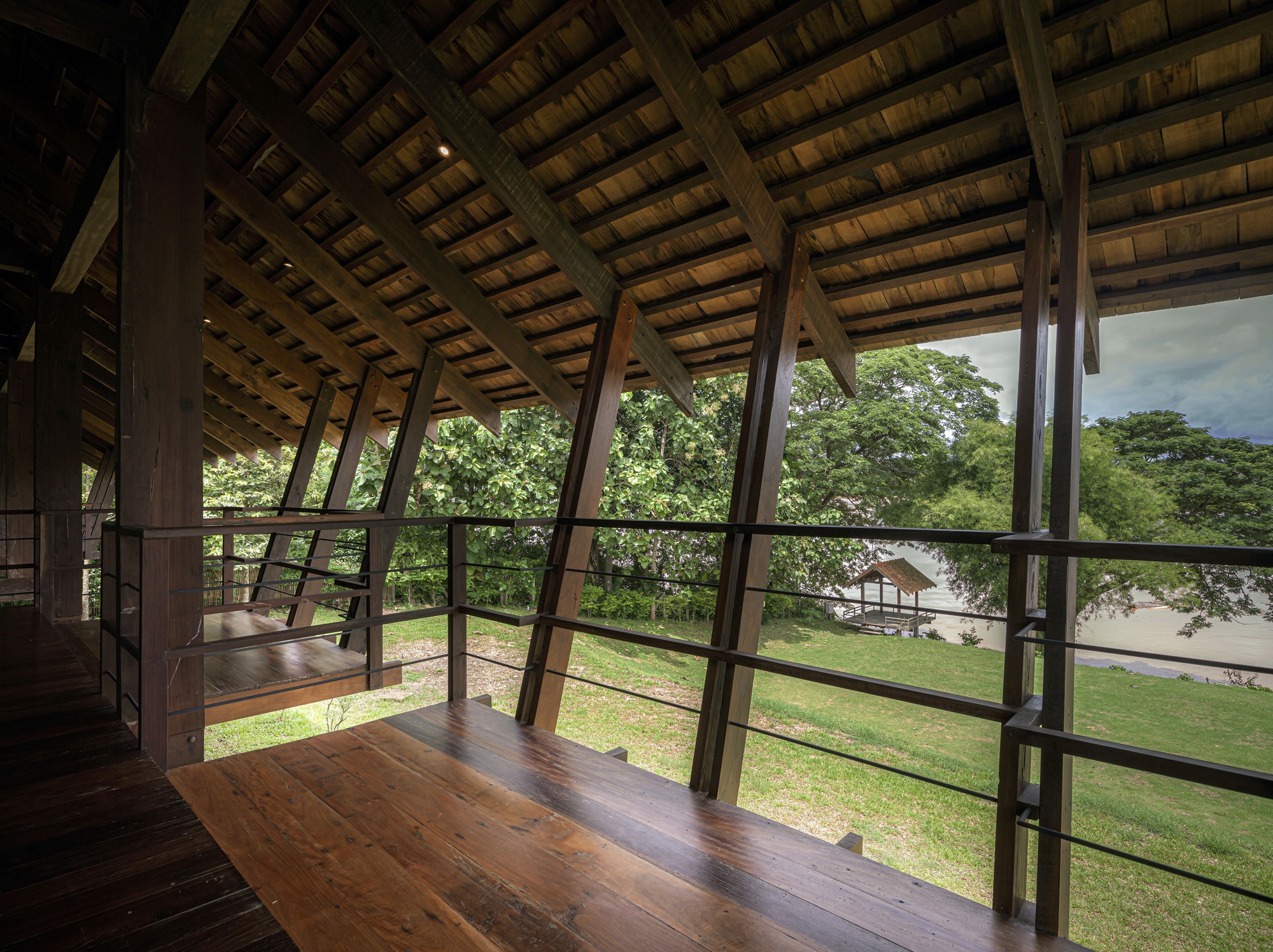 Upper floor interior with angled timber columns framing glazed walls overlooking lawn and trees beneath exposed timber ceiling