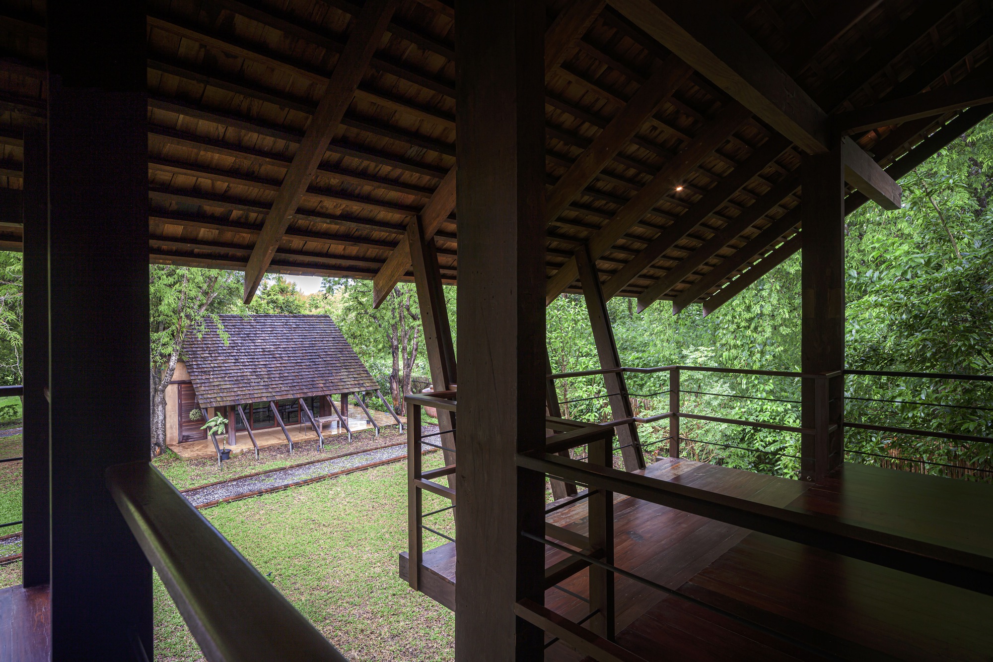 Interior looking through timber columns and steel railings to neighboring gabled structure in garden