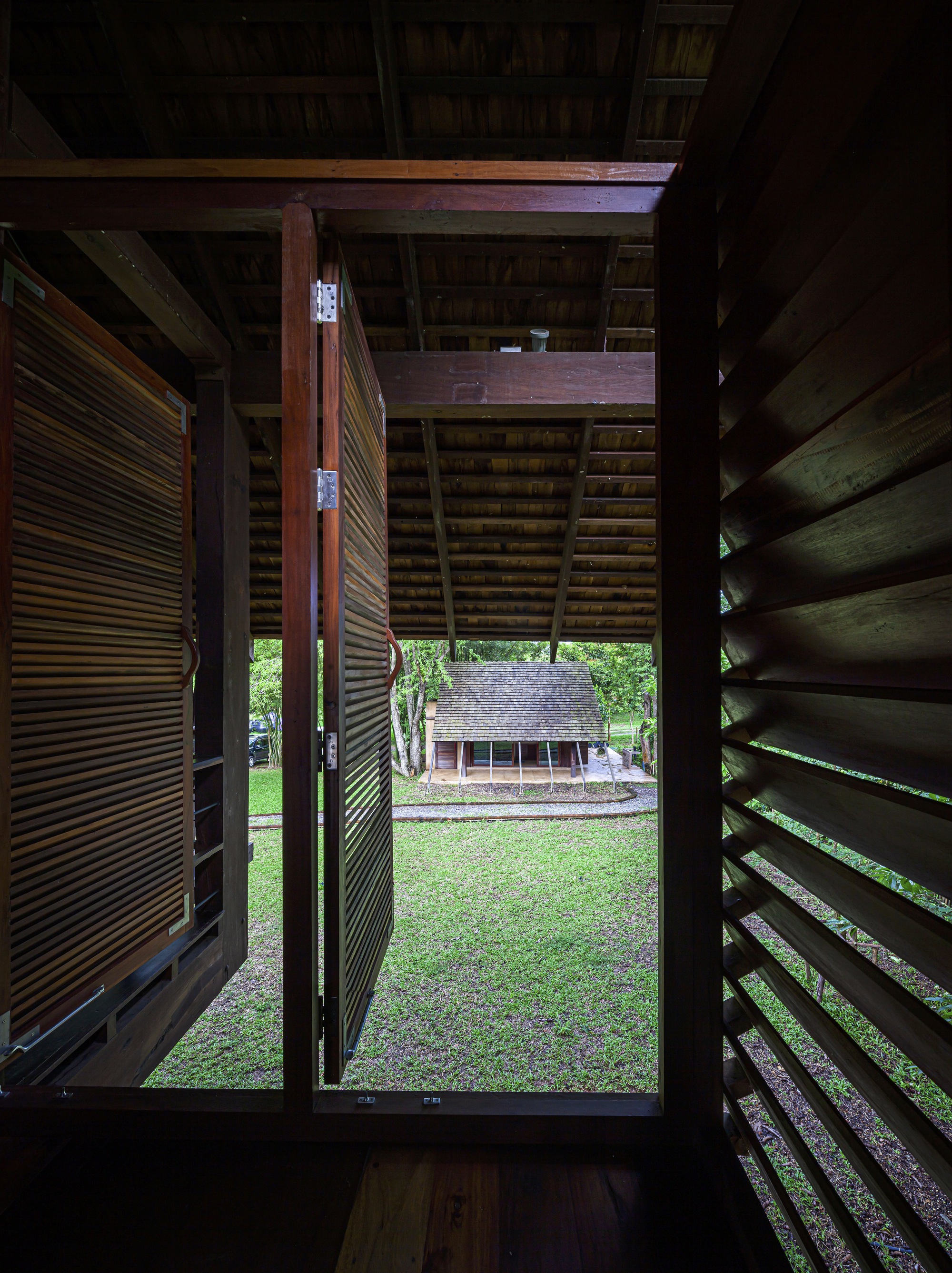 View through open timber louvered doors framing a lawn and pavilion beyond under exposed beam ceiling