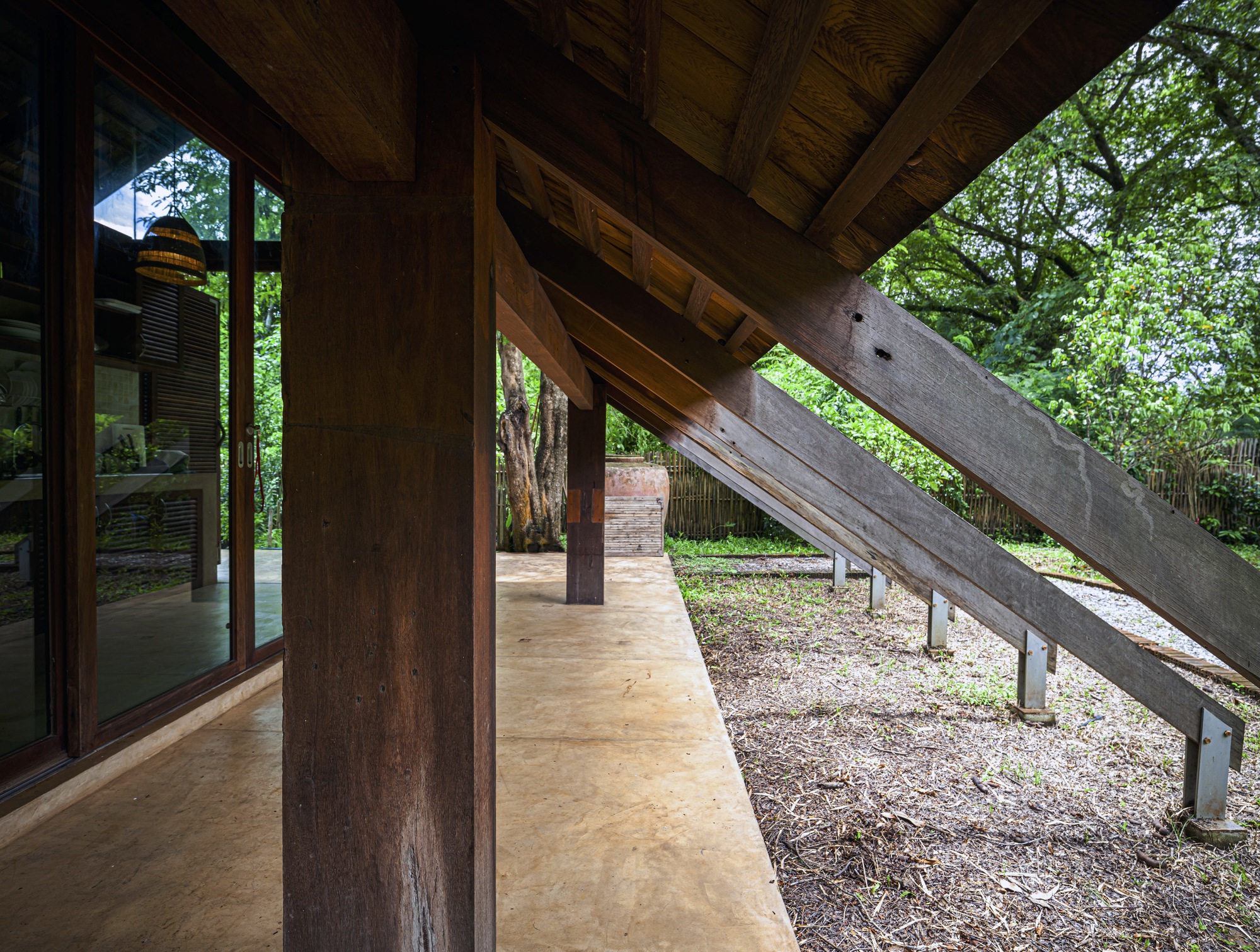 Covered terrace with timber columns and exposed beams extending toward diagonal concrete ramp and trees