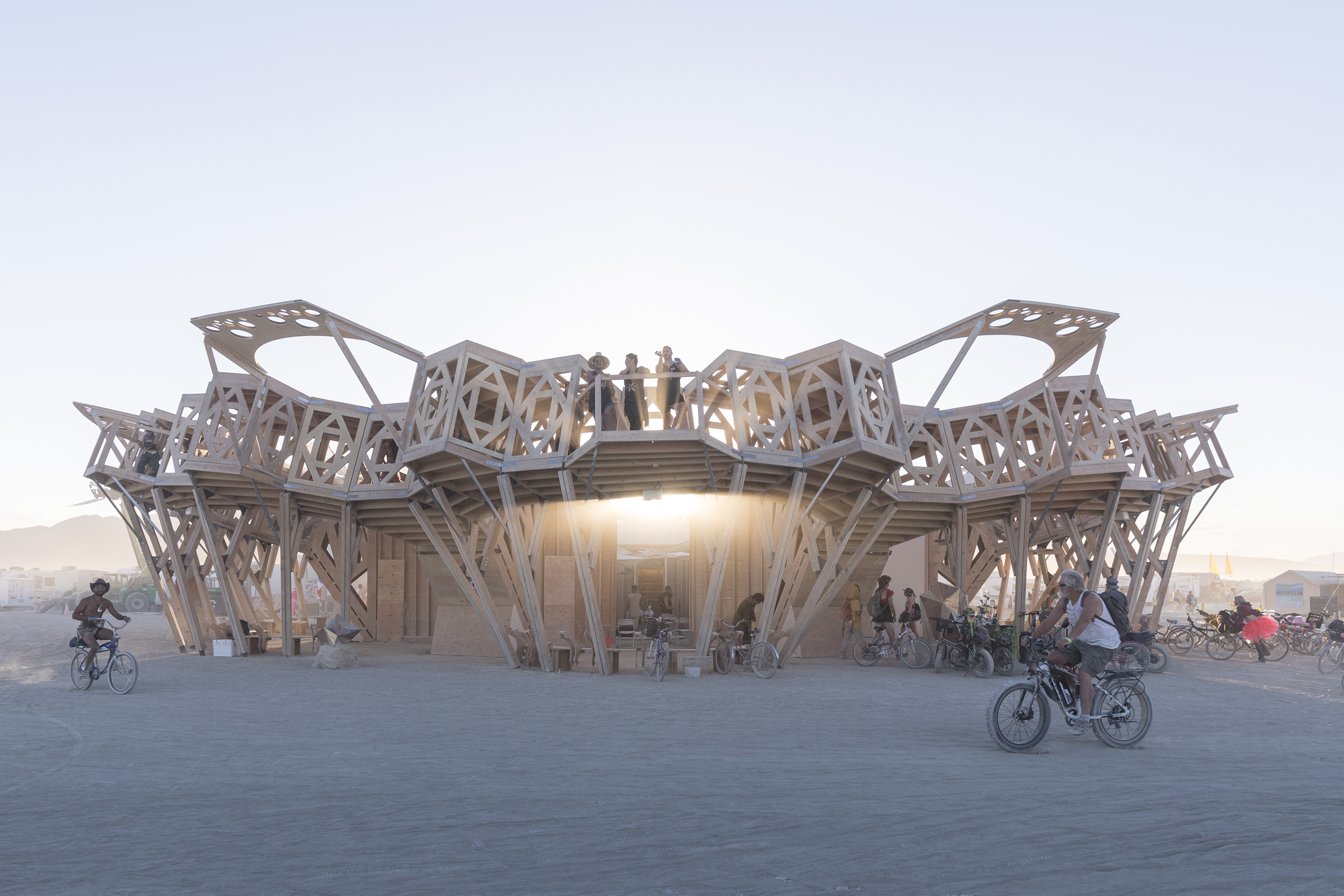 Latticed timber pavilion with patterned apertures and upper deck as cyclists pass at dusk