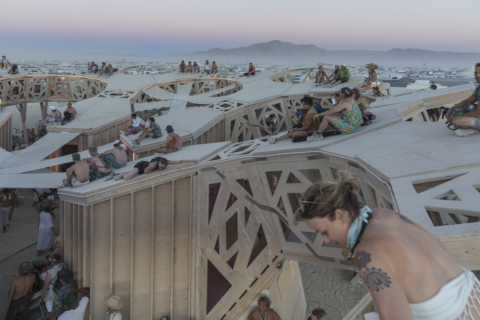 Visitors resting on tiered timber platforms with patterned parapets at twilight with mountains beyond