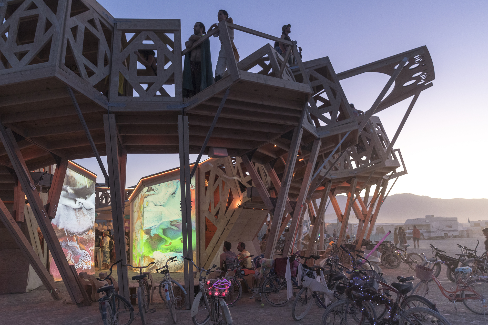Timber pavilion with open geometric lattice framework and illuminated fabric screens at dusk