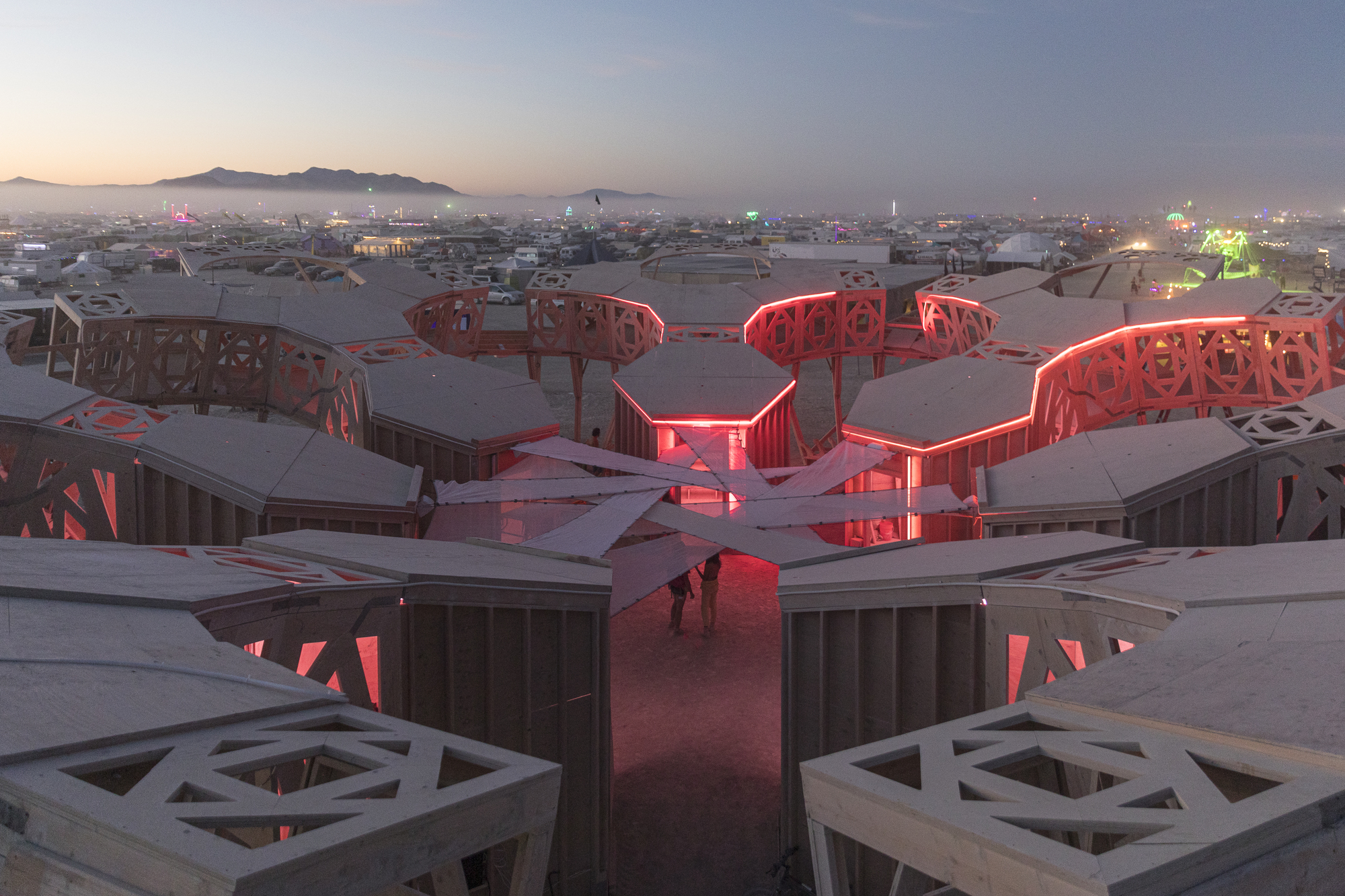 Three-lobed timber structure with red interior lighting at dusk in the desert