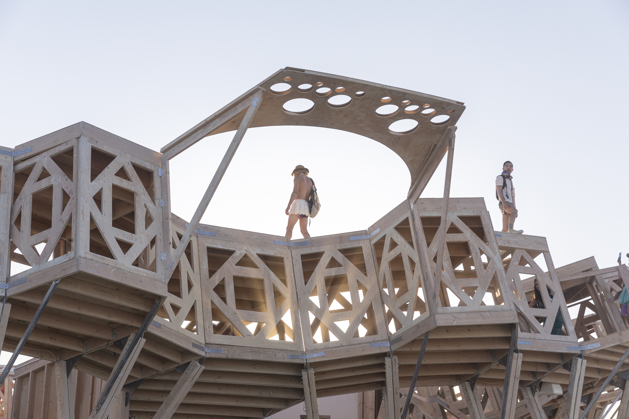 Perforated timber canopy atop geometric wooden framework with visitors silhouetted against the sky