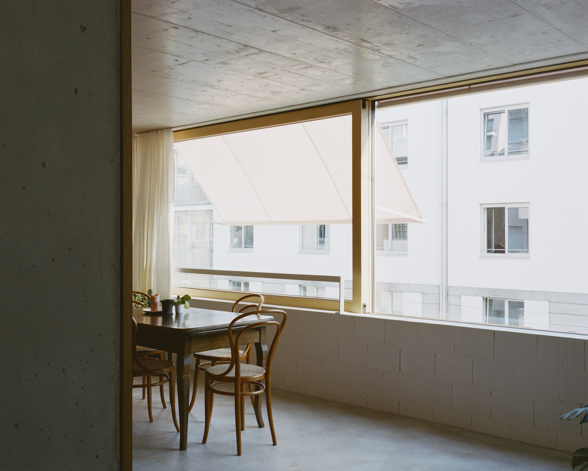 Interior corner with exposed concrete ceiling, wood-framed window and bentwood chairs at a table overlooking white facades