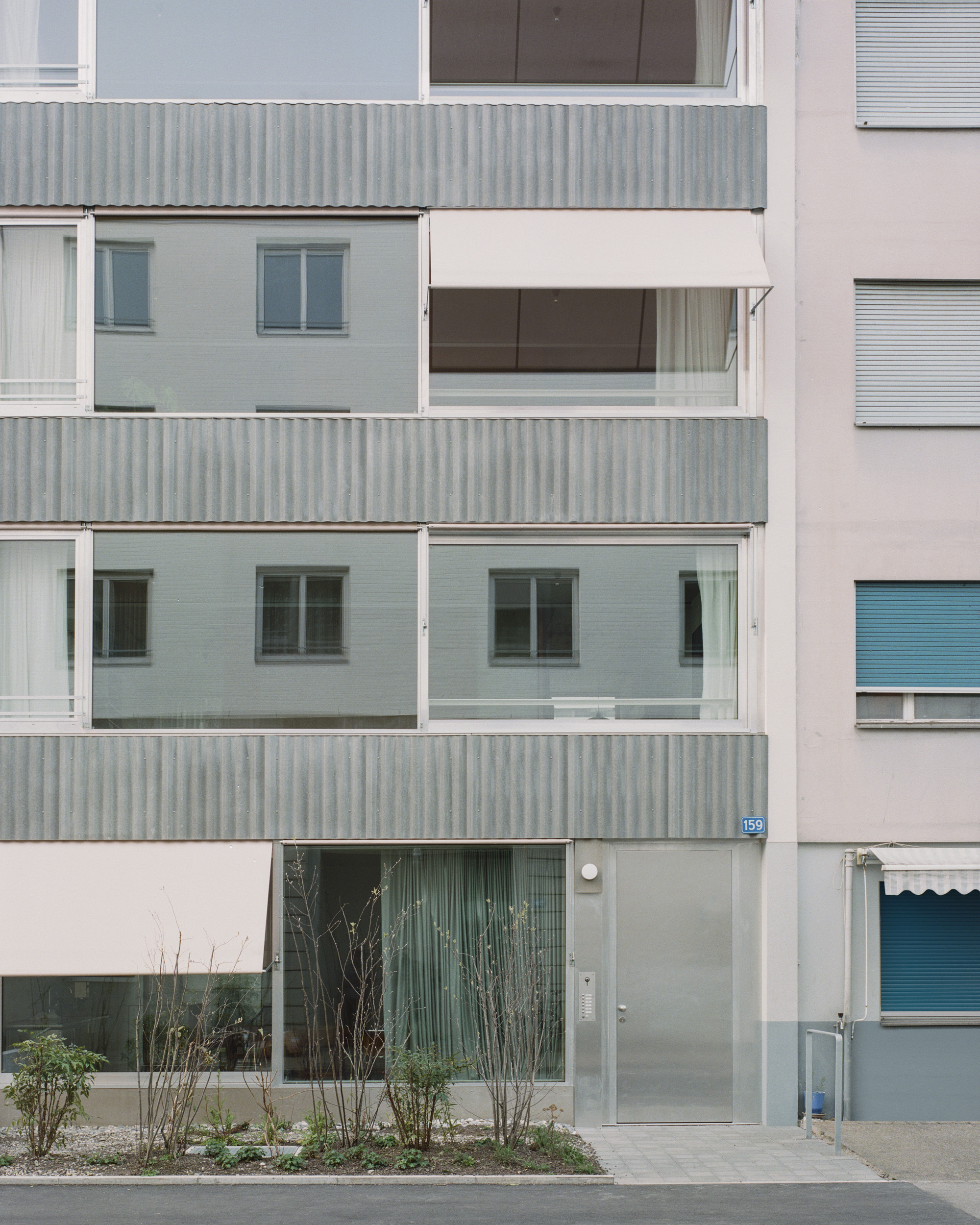Street facade with corrugated metal balconies, tiled panels and planted shrubs at ground level in overcast light