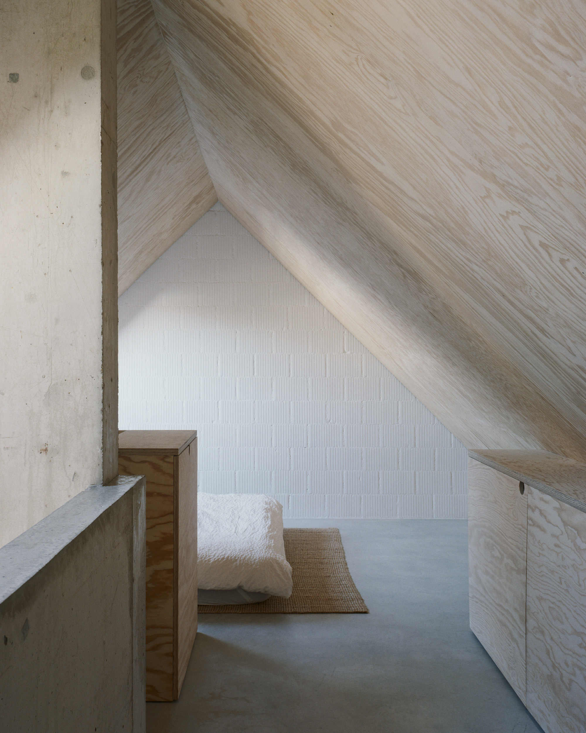 Attic space with diagonal wood-clad ceiling, concrete walls and a cushion on woven mat under soft daylight