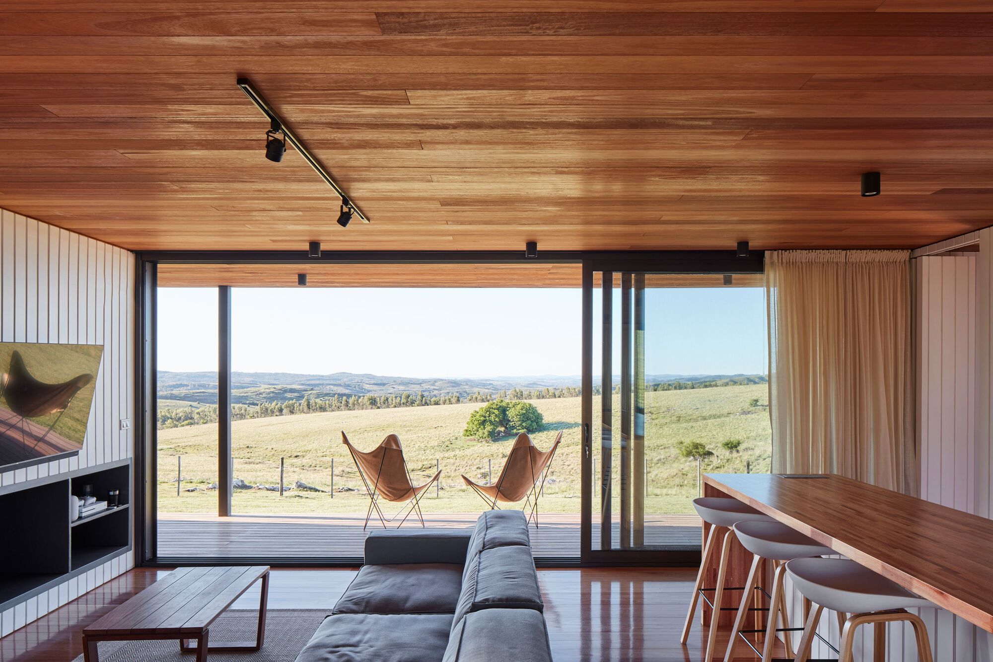 Living space with timber ceiling and full-height glazing overlooking rolling pasture and distant hills