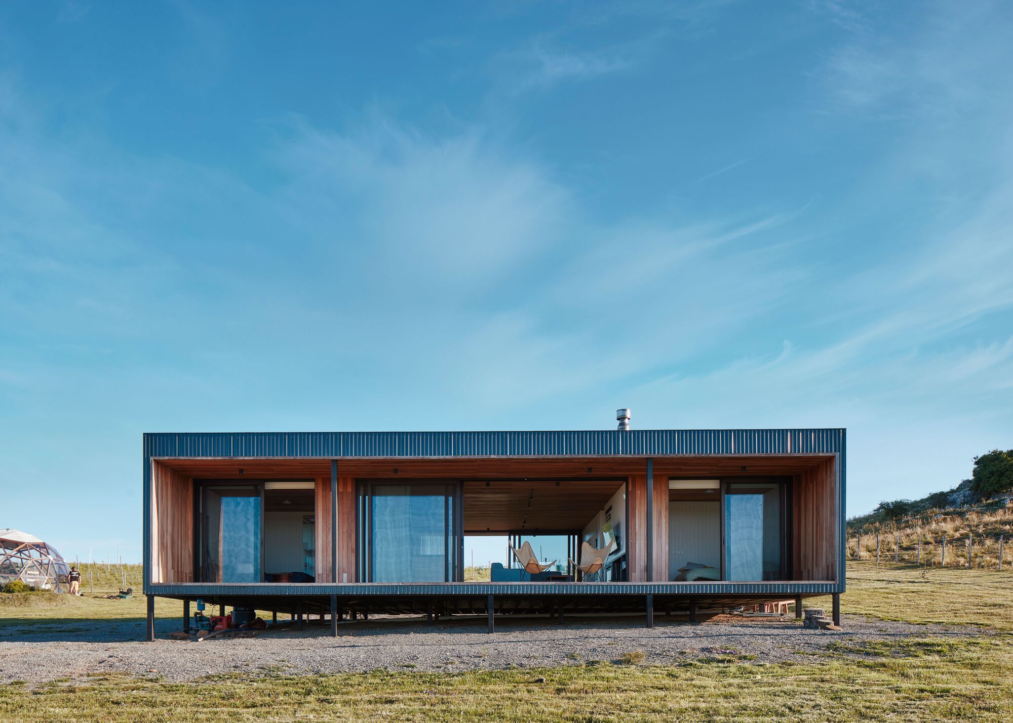 Elevated timber pavilion with full-height glazing and a corrugated metal roof on a gravel site