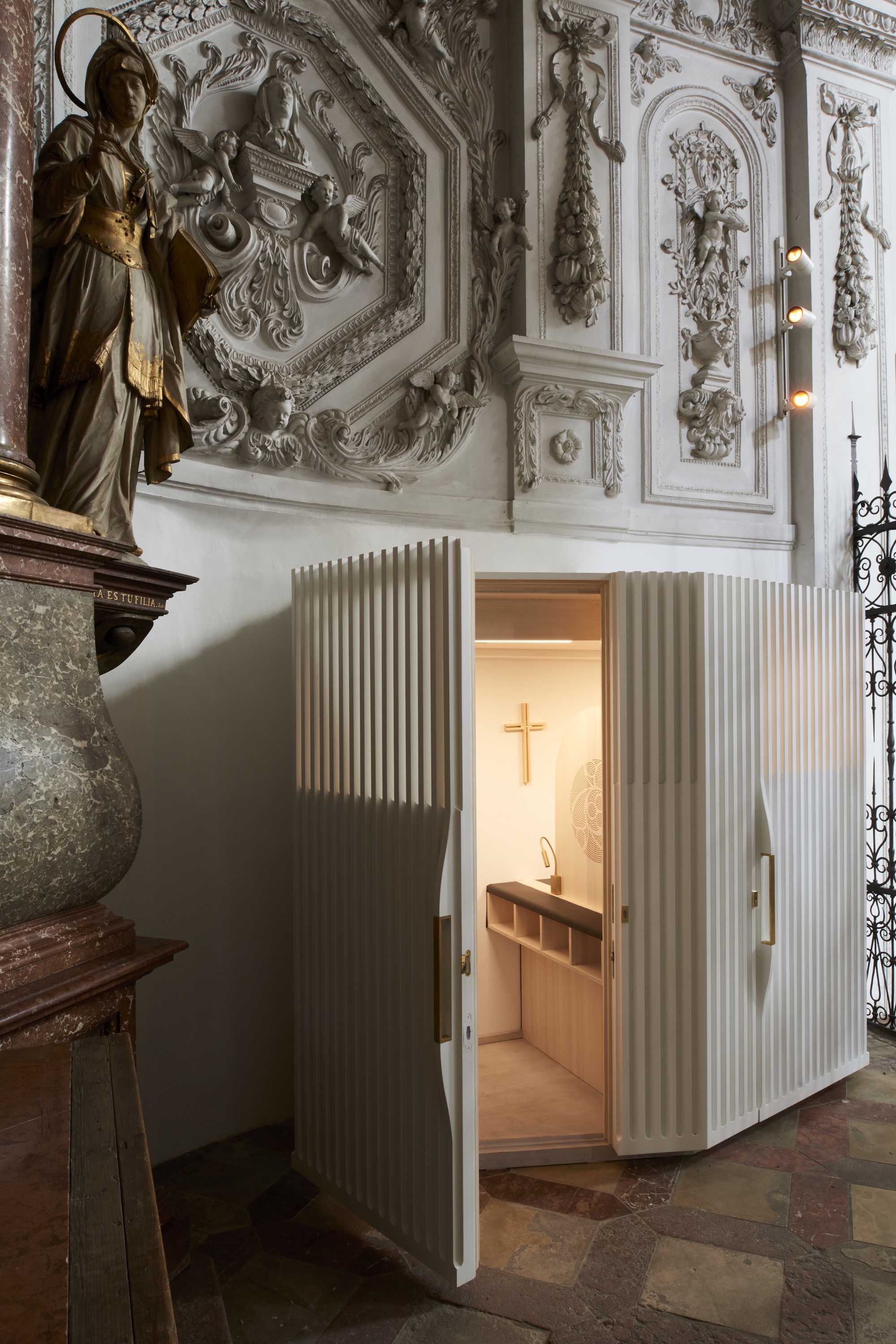 Open doorway in the ribbed timber enclosure revealing the lit confessional interior with cross and seating