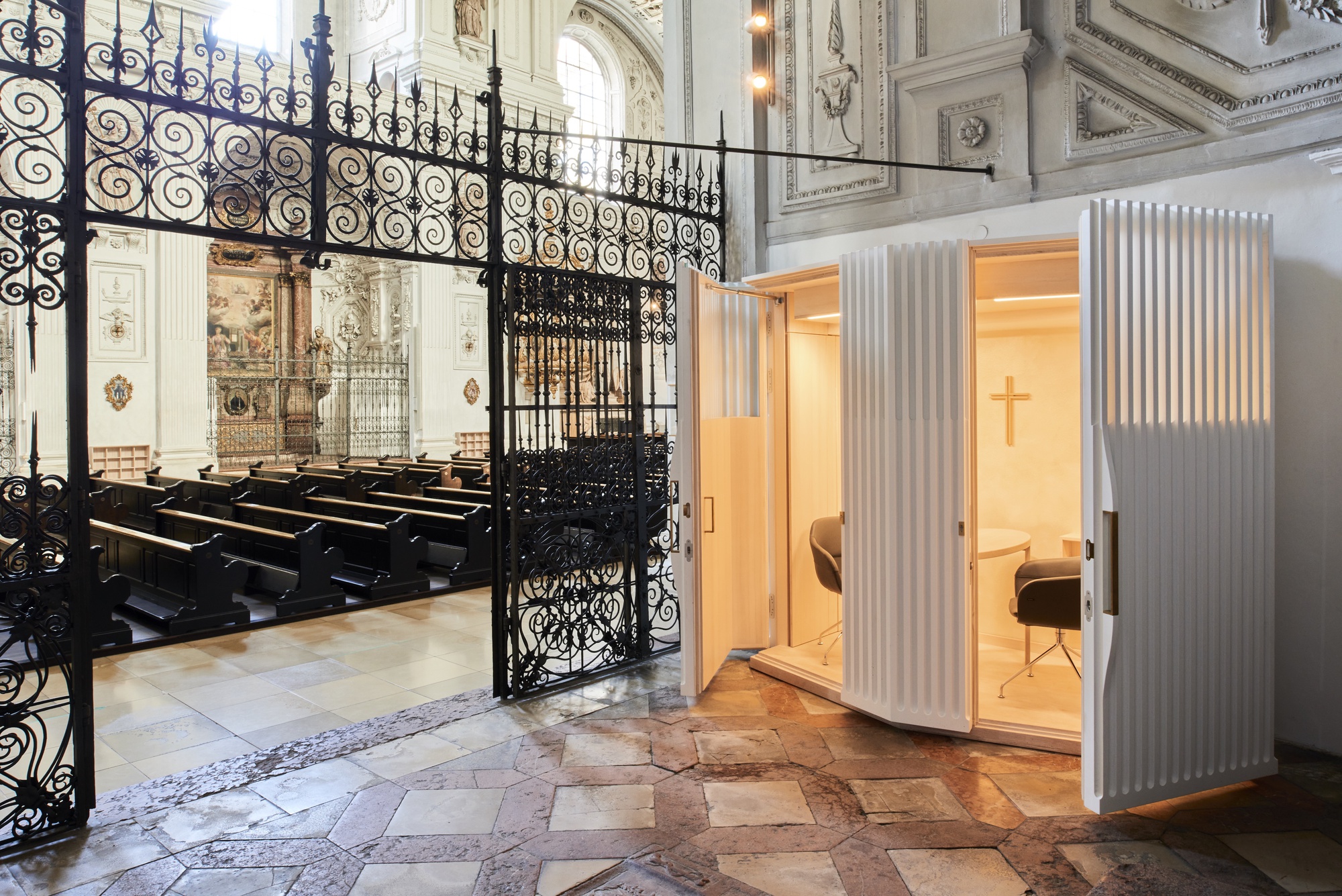White fluted confessional booth inserted within a historic church interior behind ornamental wrought iron gates