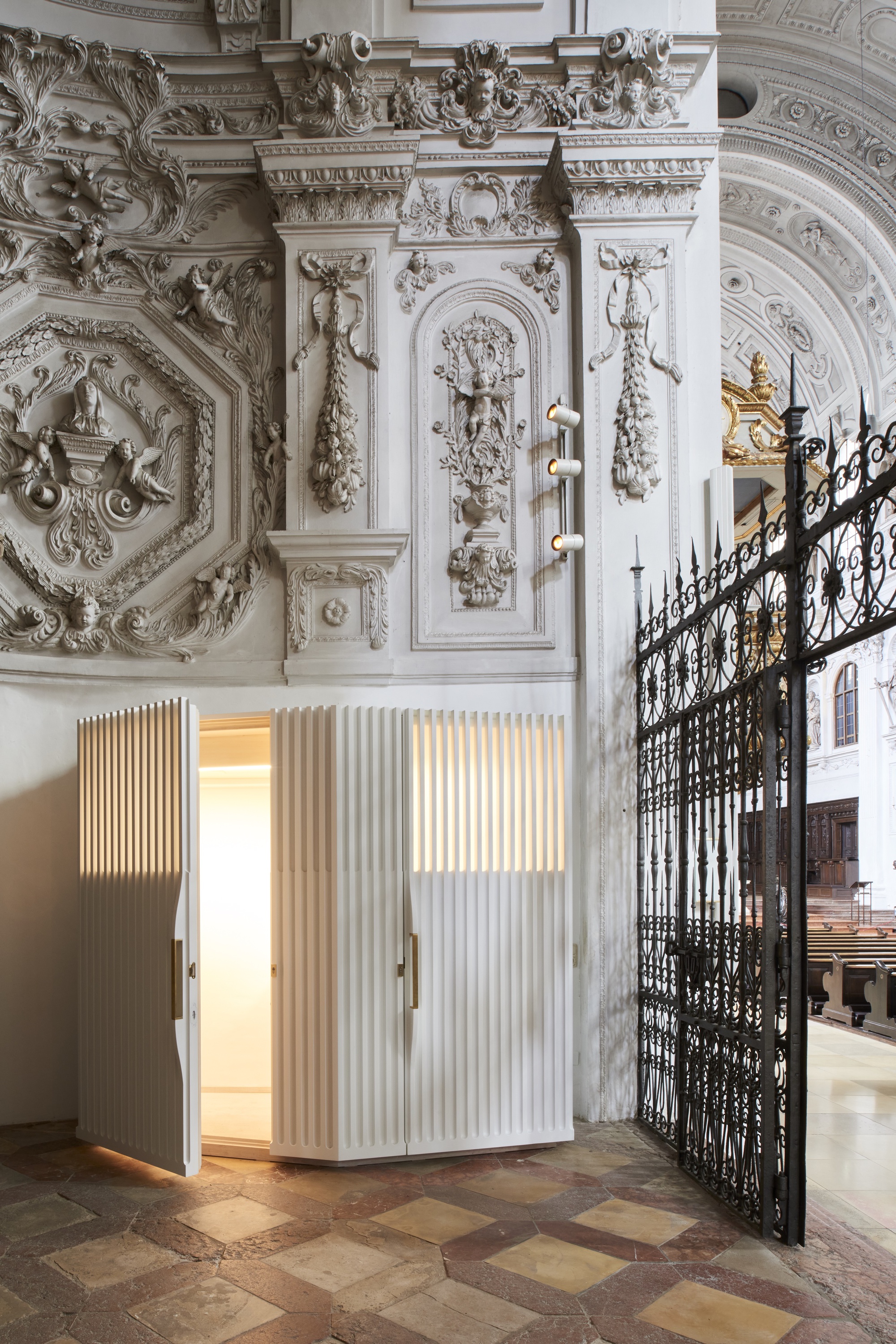 White fluted confessional booth with open door showing illuminated interior and brass handles in the church nave