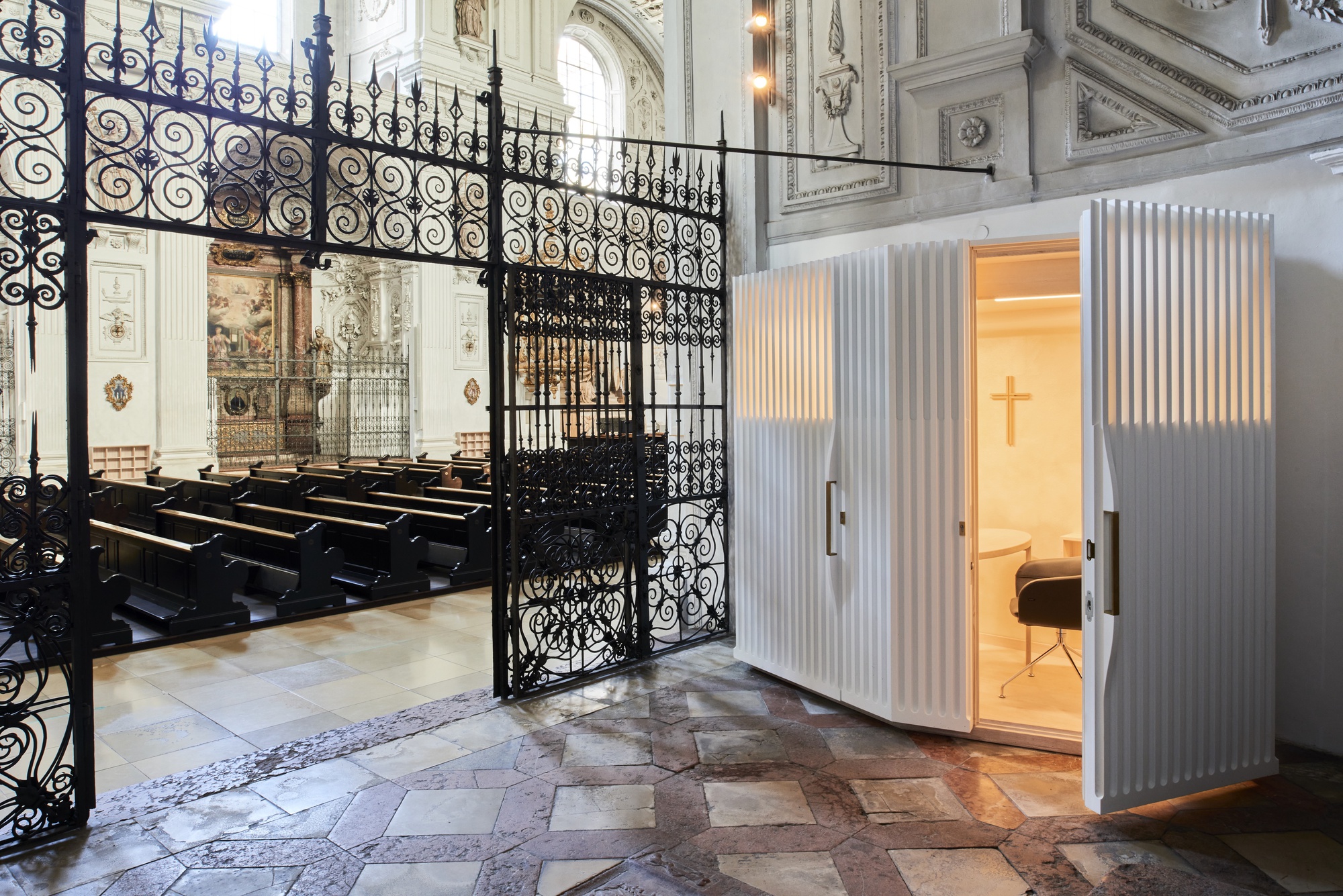 White confessional booth with illuminated cross and vertical fins beside ornate wrought iron gates and pews