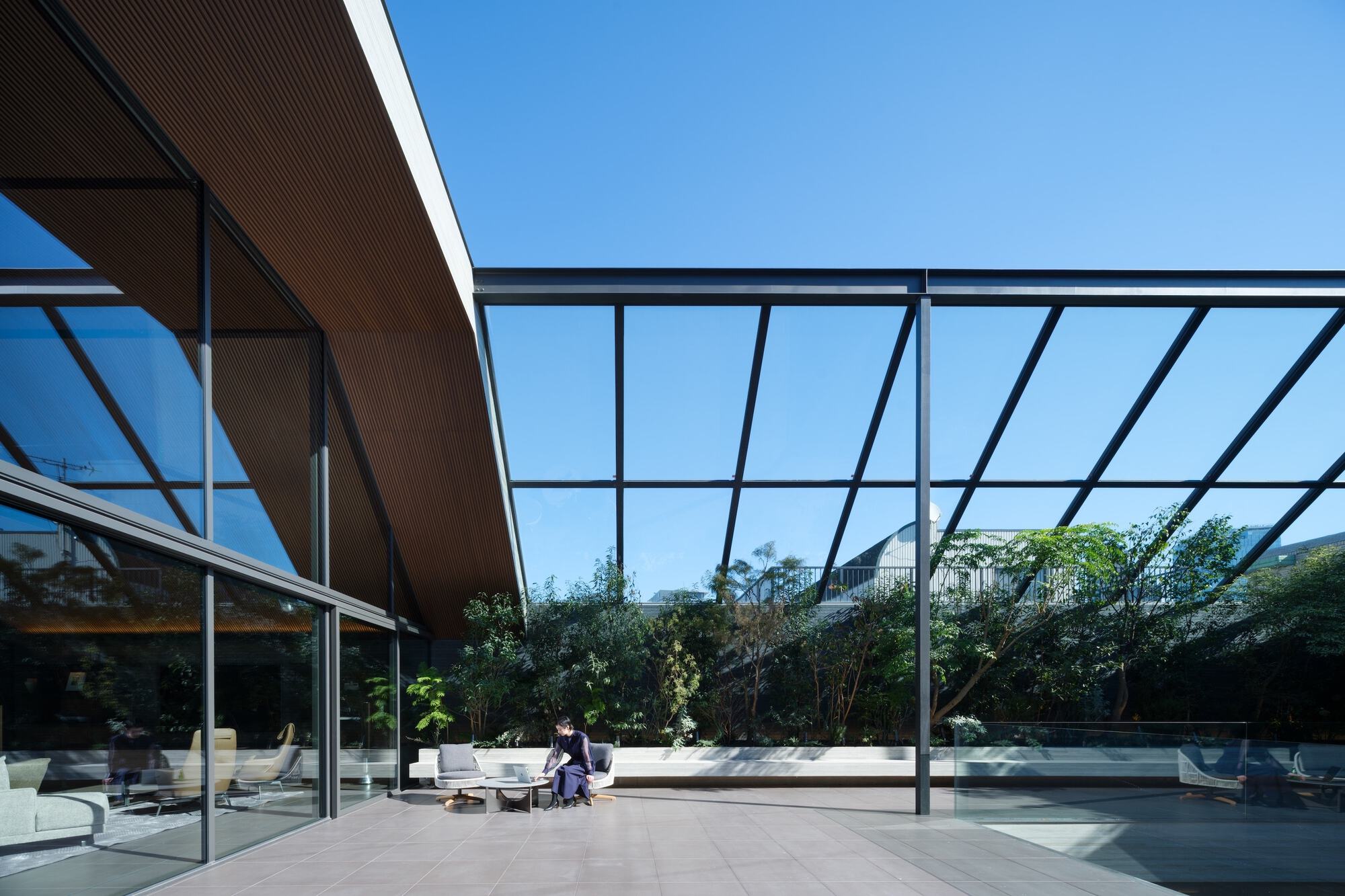 Covered terrace with steel frame and glass roof looking toward planted screening and loungers