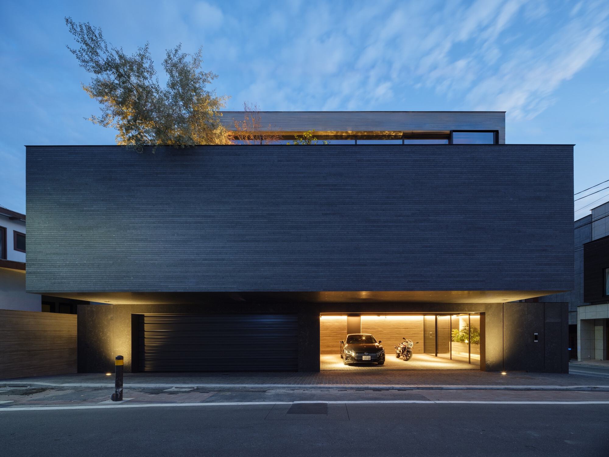 Street facade with dark brick upper volume cantilevering over an illuminated garage at twilight