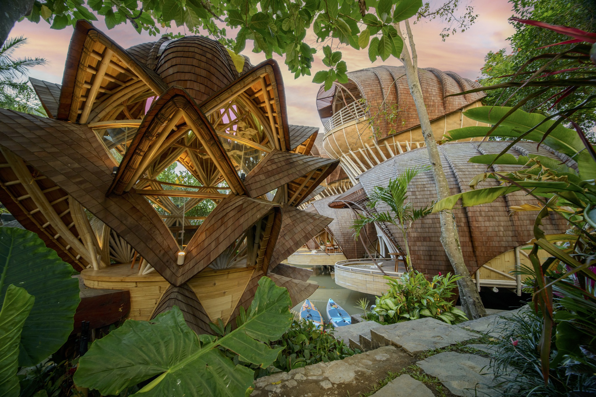 Timber lattice structure framing views of bulbous shingled volumes through dense tropical planting at dusk