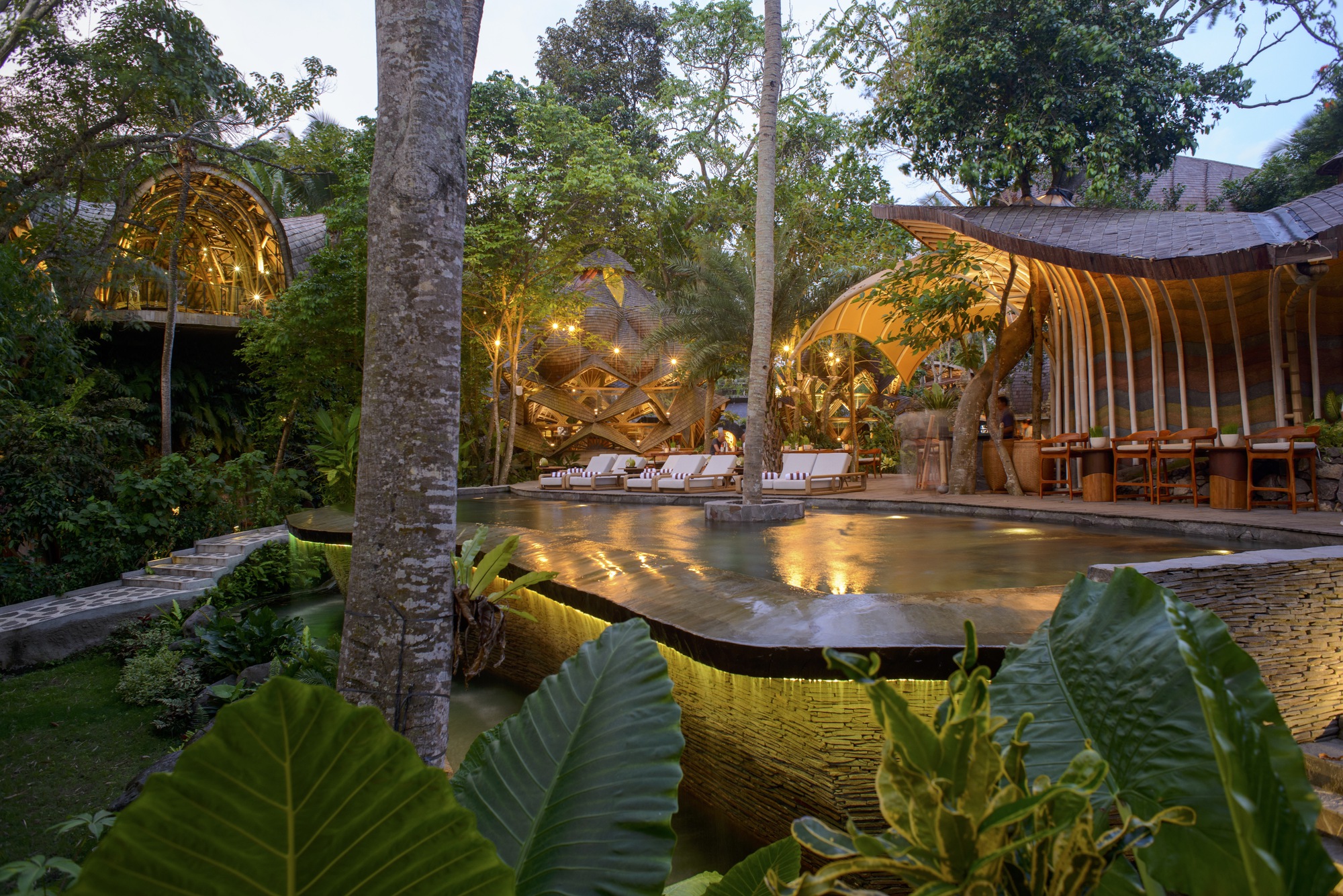 Courtyard pool at dusk framed by palm trunks and curved timber pavilions with uplighting