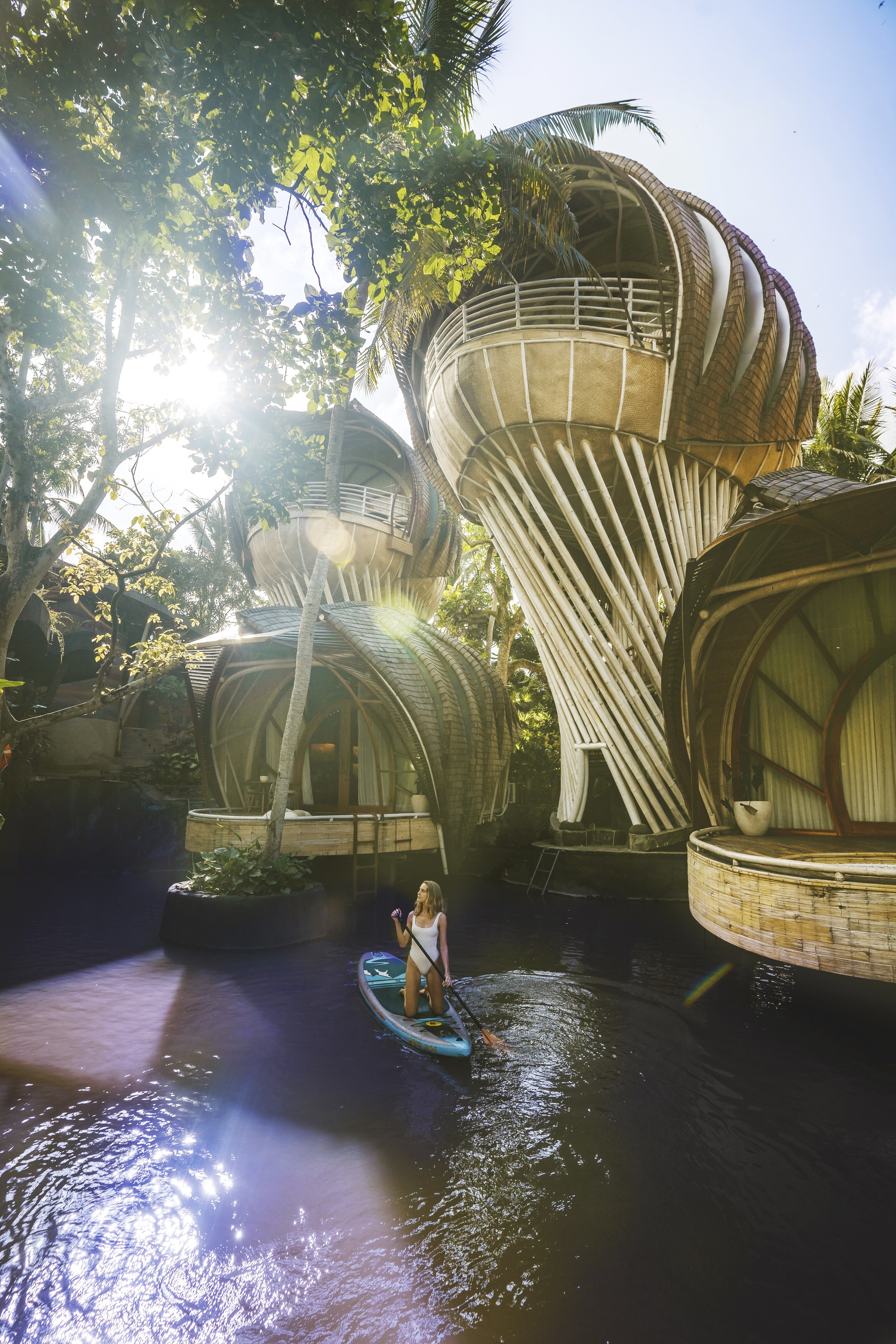 Person paddleboarding on dark water beneath clustered bamboo structures with twisted supporting columns